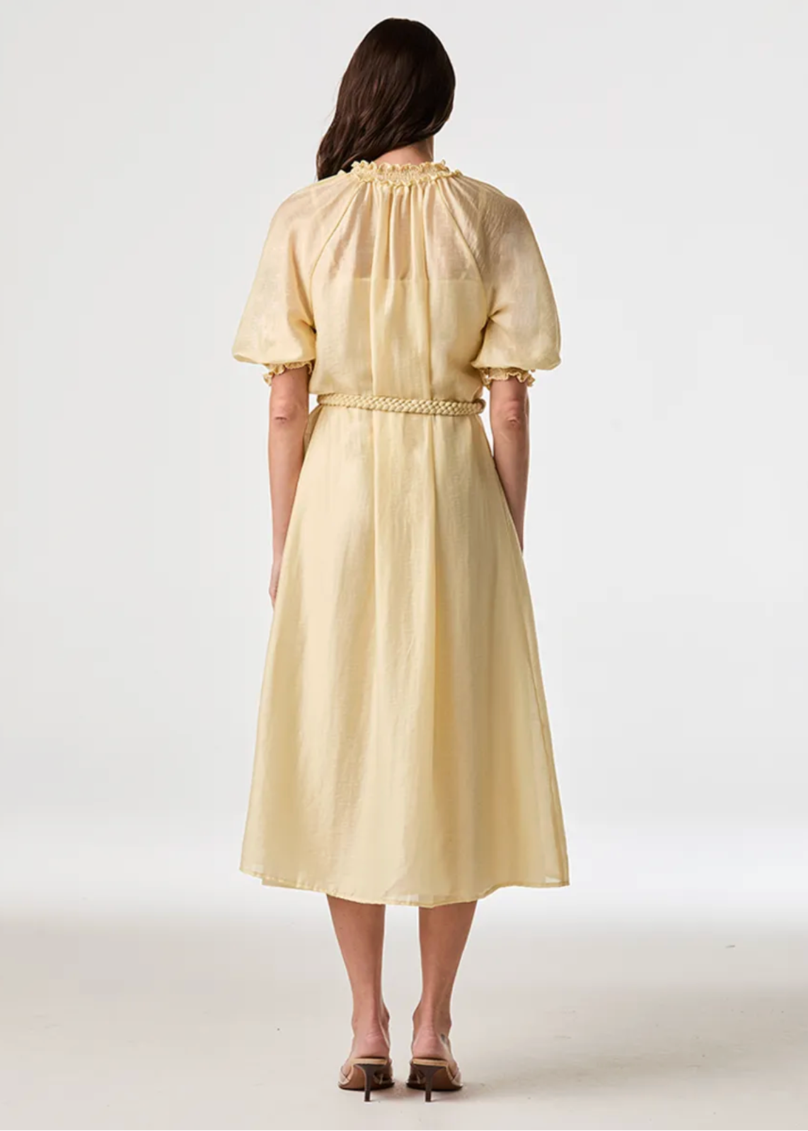 A woman viewed from the back wearing a pale yellow midi dress with short puff sleeves, a gathered neckline, and a smocked waist, photographed against a clean light gray studio background.