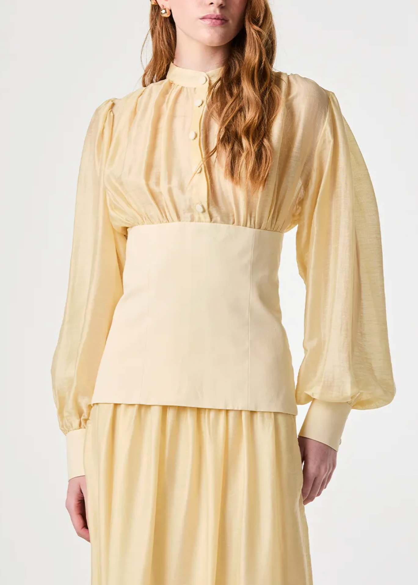 A model wearing a pale yellow long-sleeve blouse with a high mock neck, buttoned front, and a wide structured waistband, paired with a matching skirt against a plain light background.