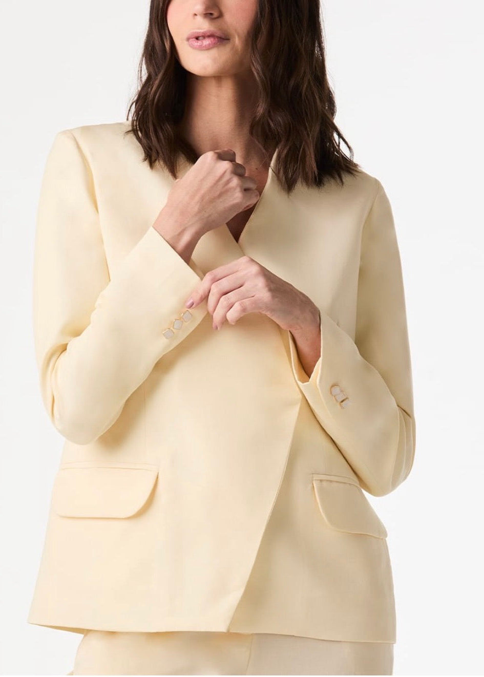 A woman wearing a pale cream or light beige tailored blazer with structured shoulders, flap chest pockets, and decorative gold-toned buttons on the cuffs, photographed against a clean light gray studio background.