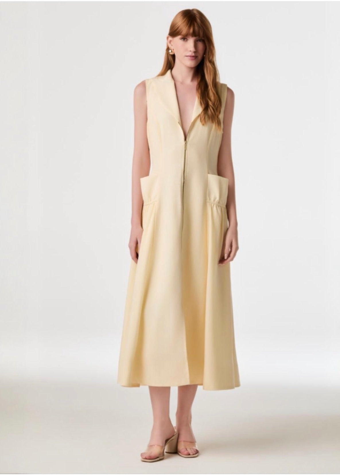 A woman modeling a sleeveless, V-neck midi dress in a pale pastel yellow, standing against a plain light gray studio background.