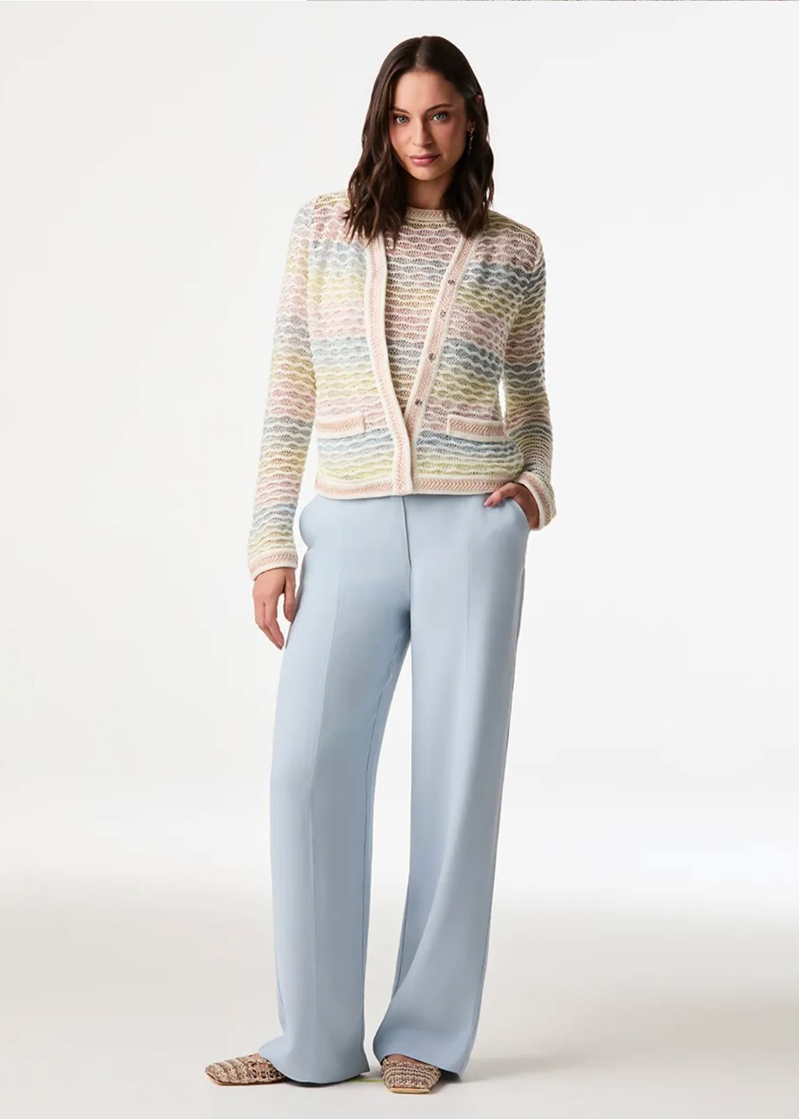 A model stands against a plain light gray studio background wearing a pastel multicolor knit zip-front cardigan over a matching knit top, paired with light blue wide-leg pants and flat woven sandals.