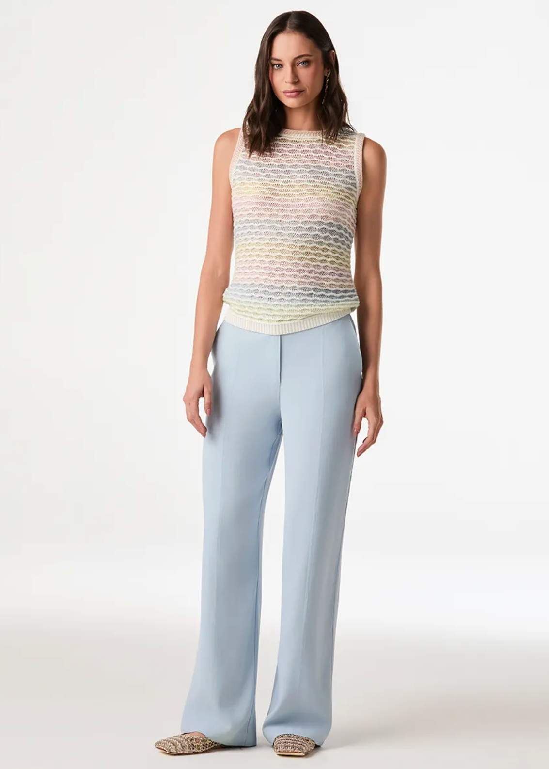 A model stands against a clean white studio background wearing a sleeveless knit top with a pastel rainbow gradient and light blue wide-leg trousers.