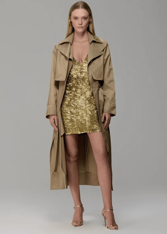 Woman wearing a gold dress and beige trench coat on a plain background