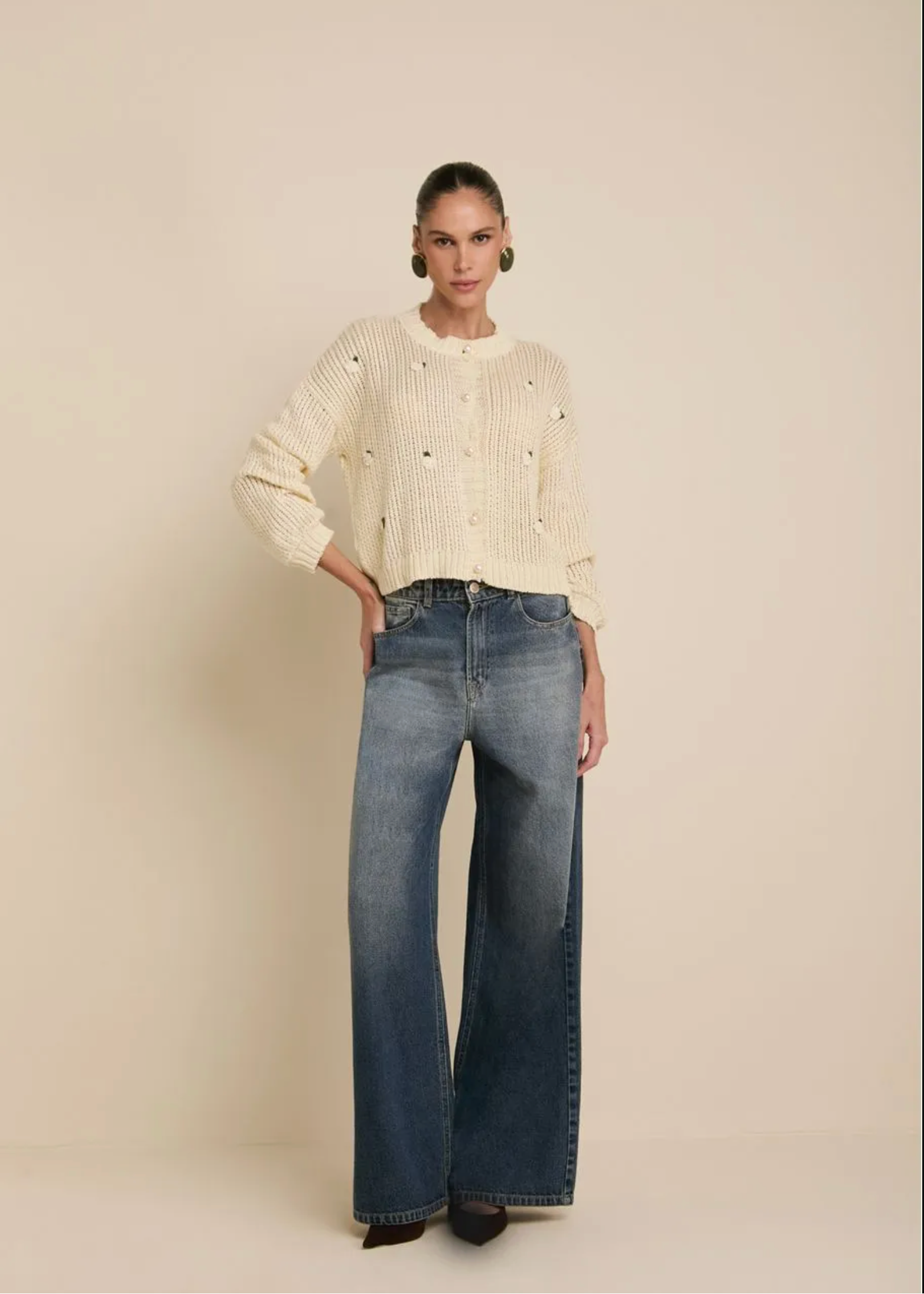 A model stands against a plain beige studio background wearing a cream-colored textured knit sweater with small black embroidered details, paired with wide-leg blue jeans.