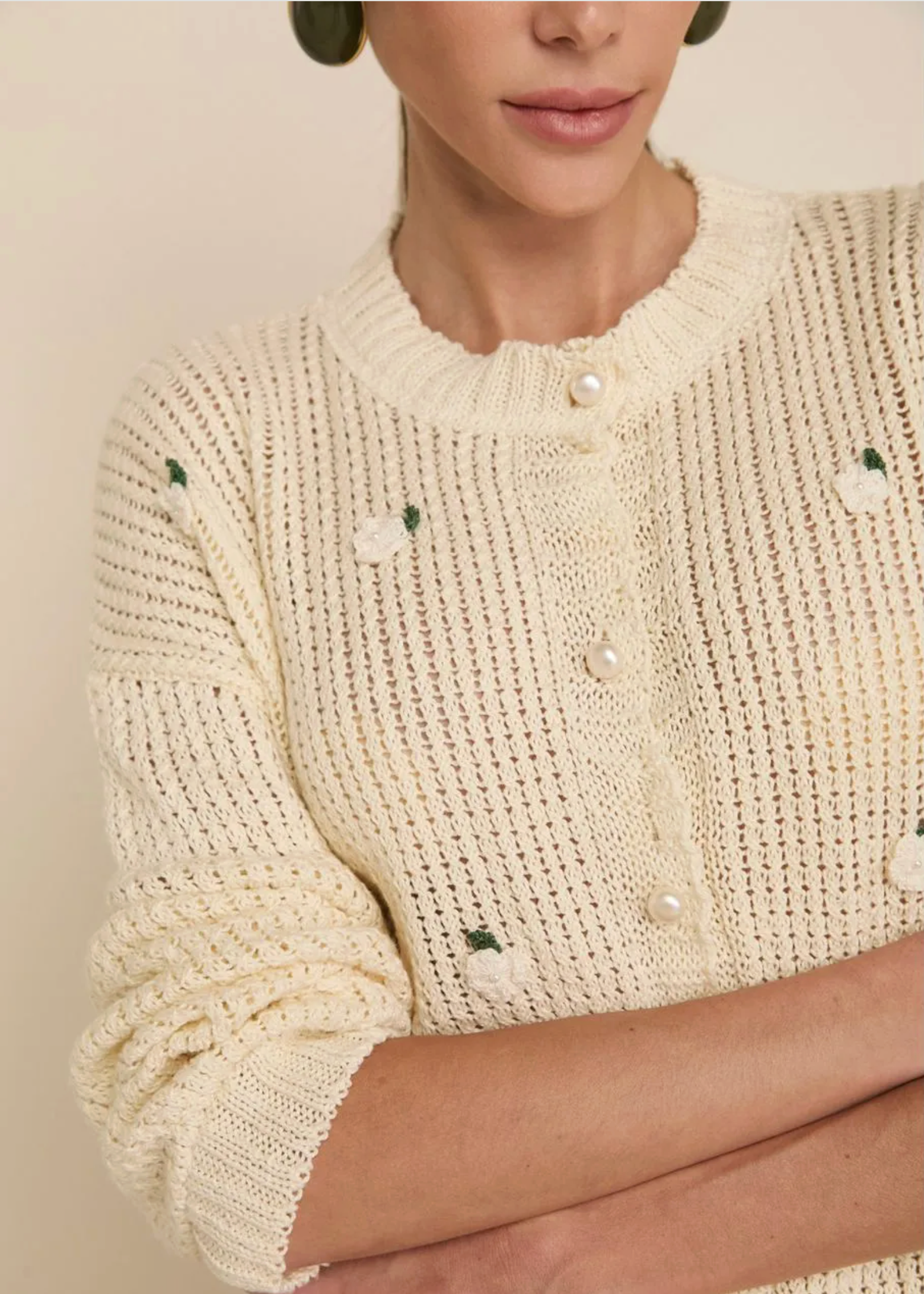 A close-up of a woman wearing a cream-colored knit cardigan with small white and green embroidered floral details and pearl-like buttons. The background is a solid, warm beige tone.