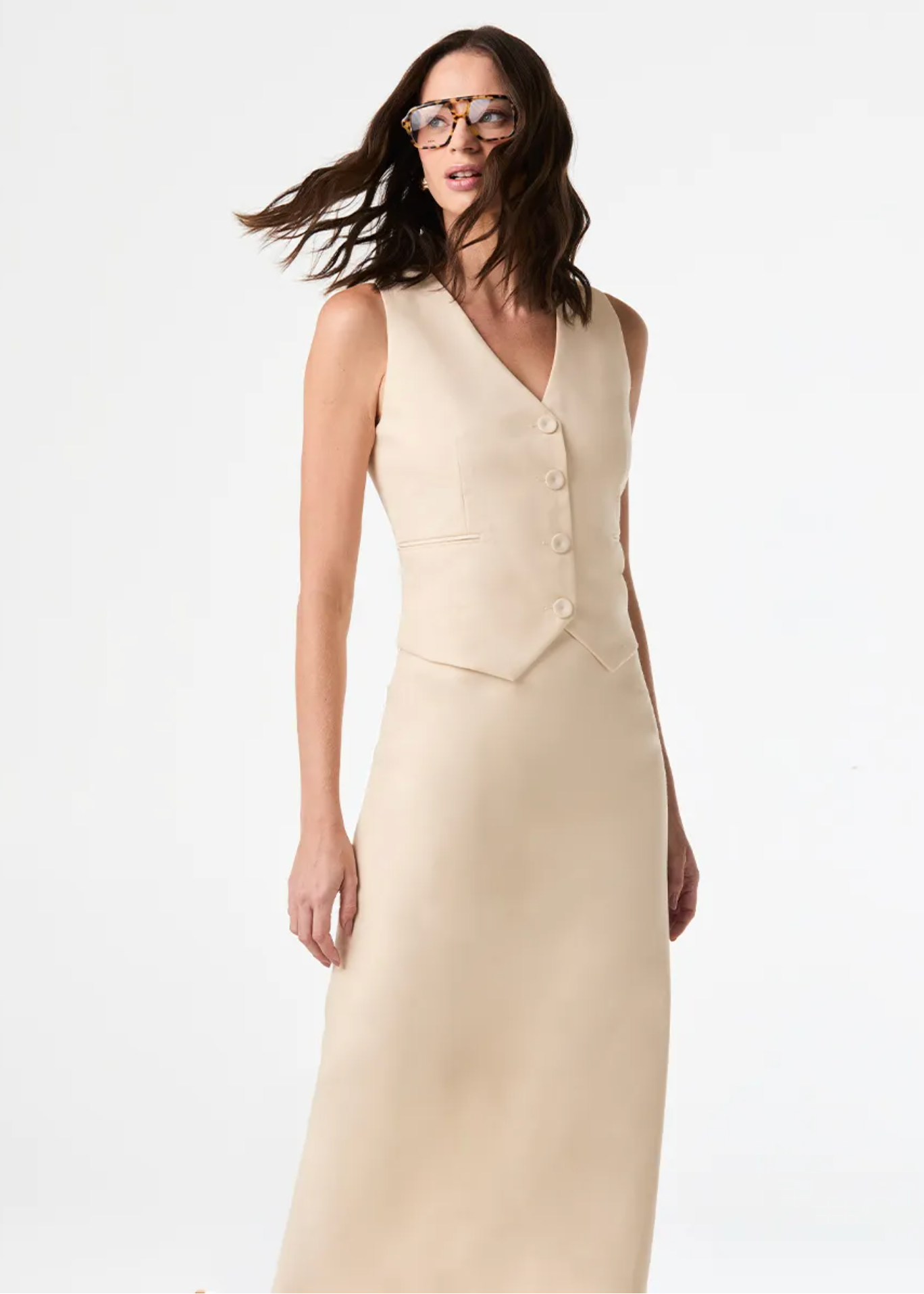 A woman modeling a sleeveless, V-neck, button-front midi dress in a light beige or cream color, set against a plain white studio background.