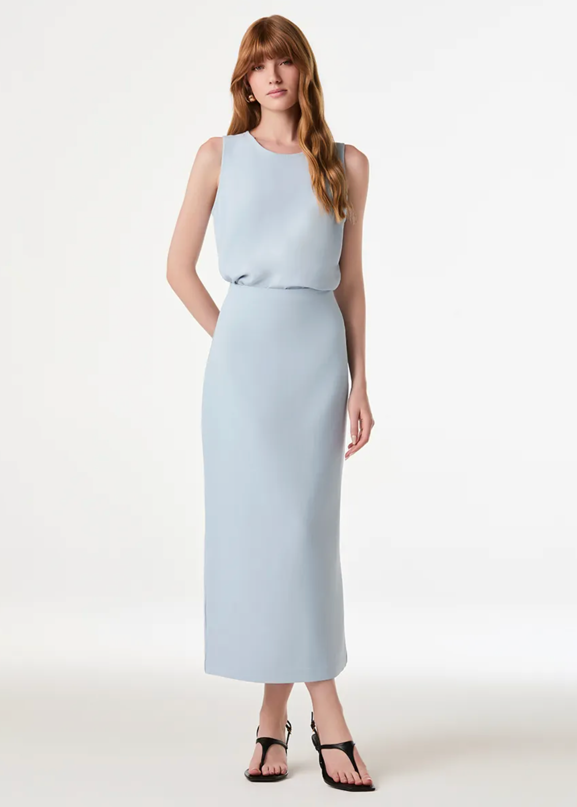 A woman modeling a sleeveless light blue midi dress with a structured bodice and a straight skirt, paired with black strappy sandals, against a plain white studio background.