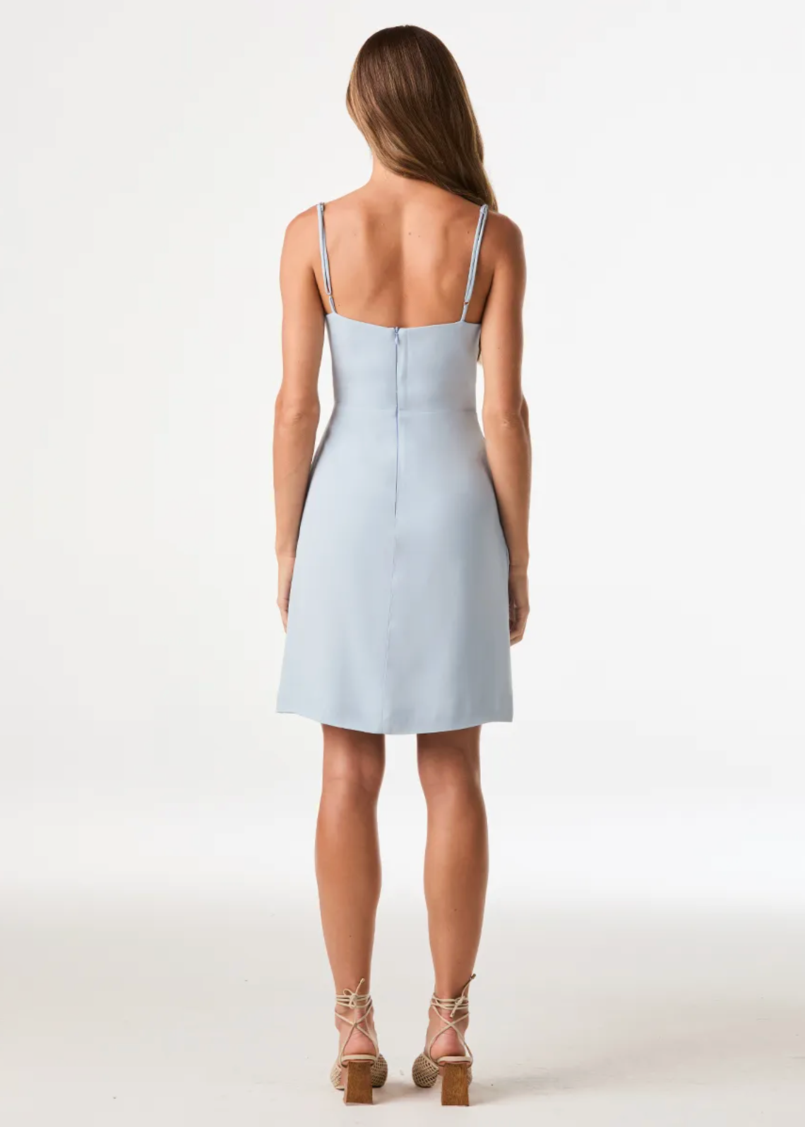 A woman viewed from the back wearing a light blue sleeveless mini dress with thin straps and a straight back zipper, paired with beige lace-up heeled sandals, against a plain white studio background.