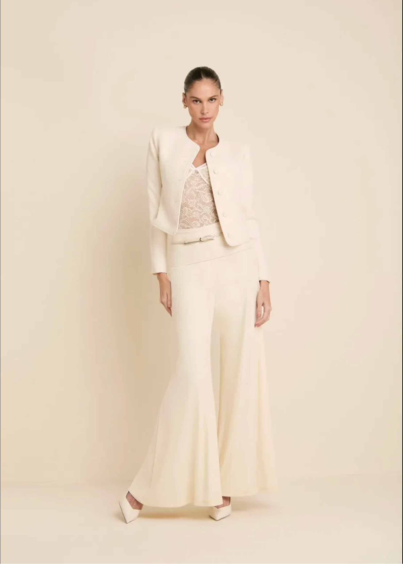 A model stands against a warm cream studio backdrop wearing a matching cream two-piece suit with a cropped jacket, wide-leg trousers, and a slim belt over a lace-trimmed top.