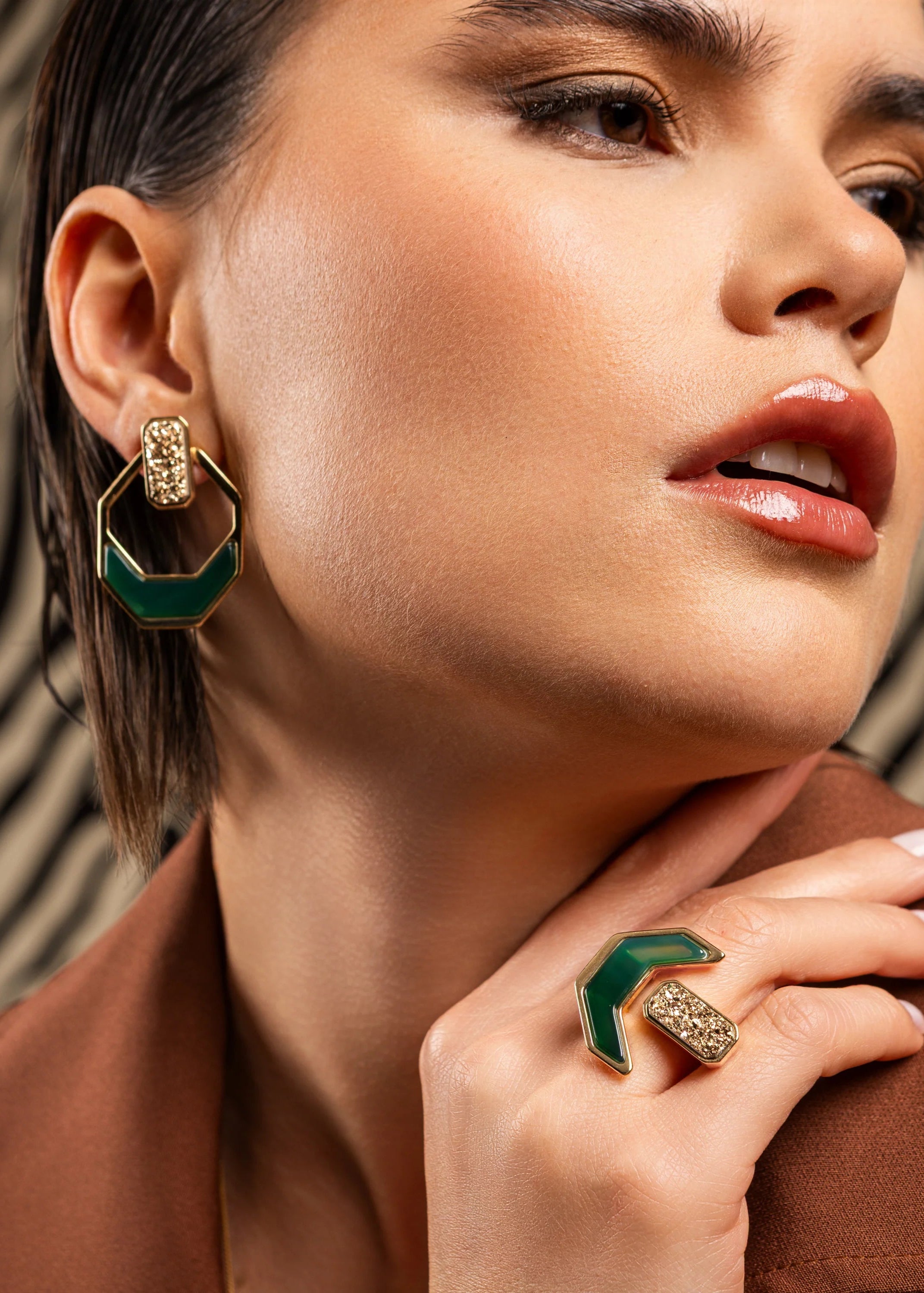 Close-up of a woman wearing green and gold earrings and ring against a patterned background
