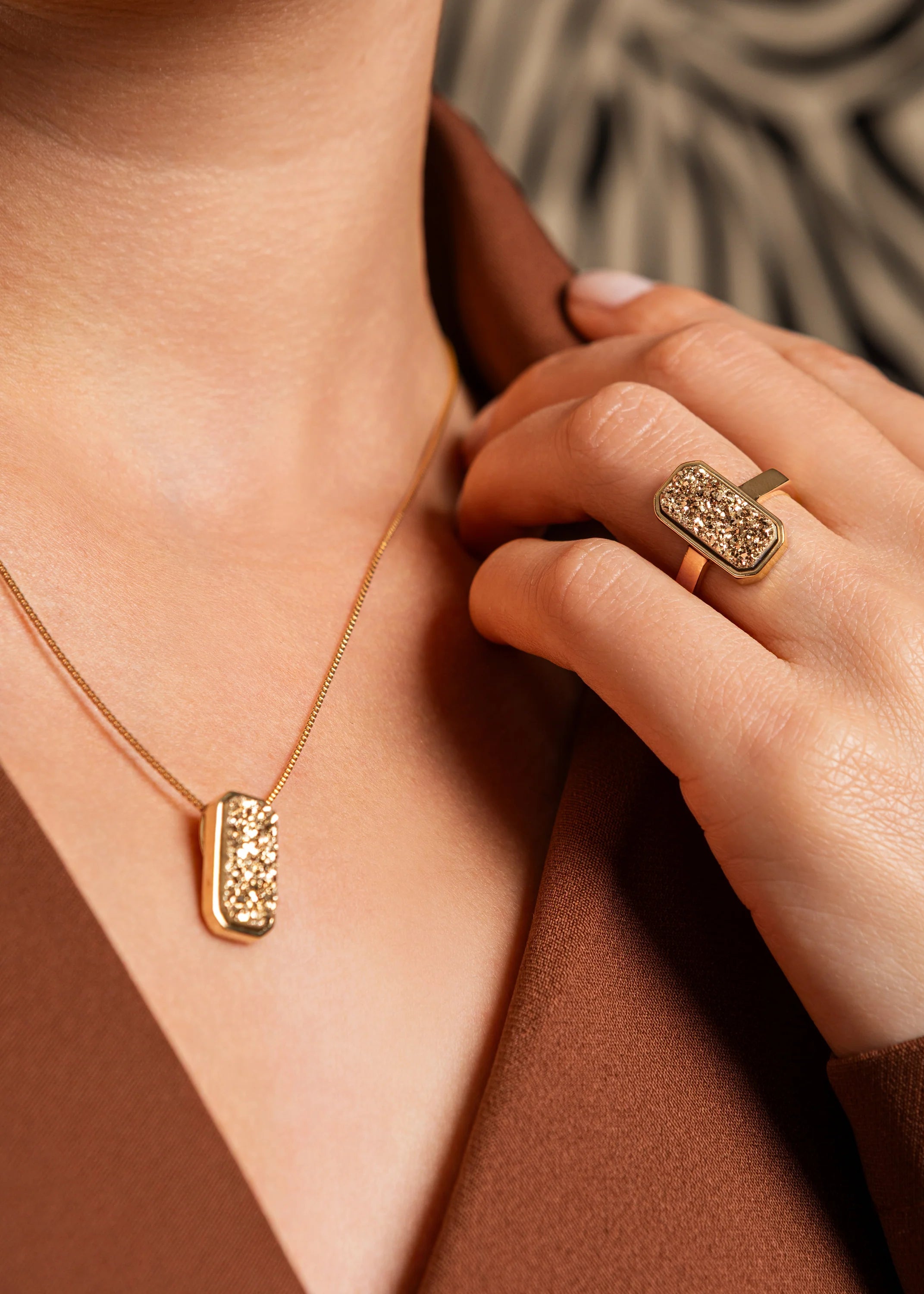 Gold necklace and ring on a person's hand and neck against a brown background
