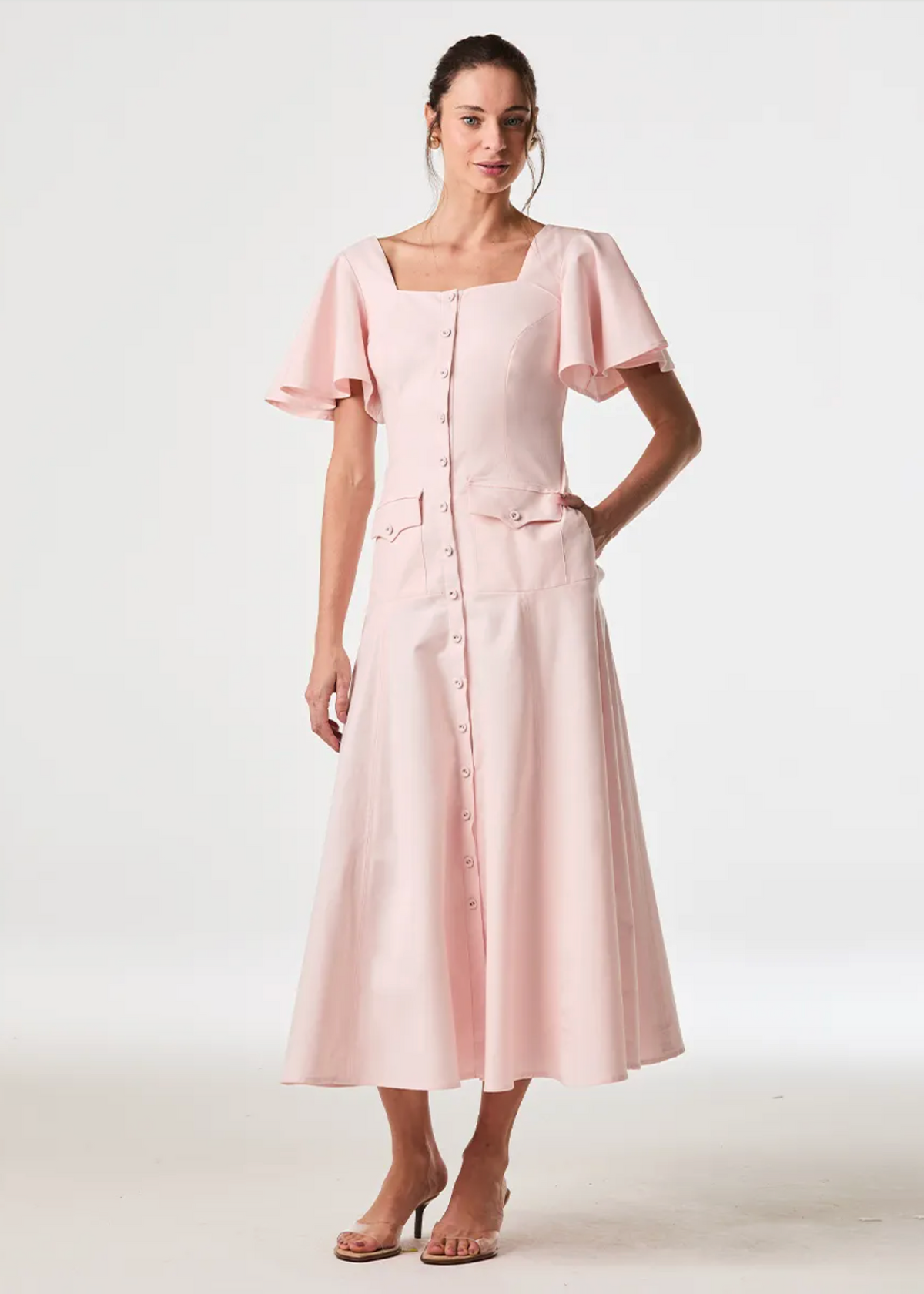 A woman modeling a light pink midi dress with a square neckline, flutter sleeves, a front button placket, and side pockets, photographed against a clean white studio background.