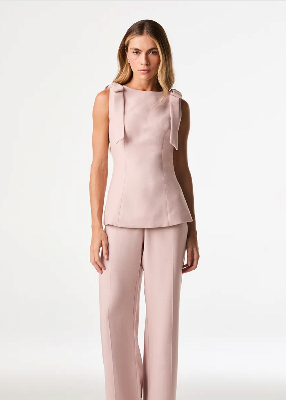 A model stands against a plain light pink studio background wearing a pale blush pink sleeveless top with bow details at the shoulders and matching wide-leg trousers.
