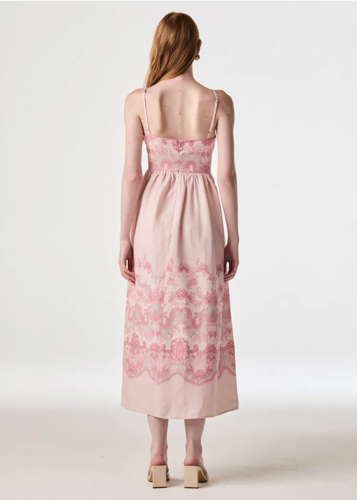 A woman stands with her back to the camera wearing a light pink midi dress with thin straps and pink floral lace detailing along the neckline and lower skirt, photographed against a plain light gray studio background.