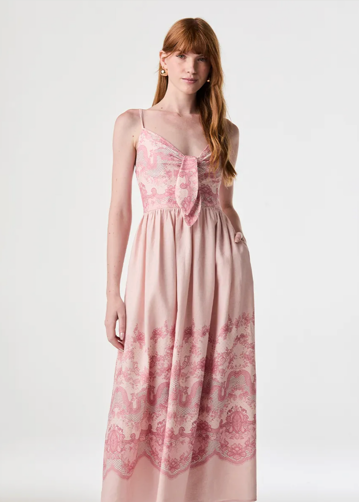 A woman modeling a light pink maxi dress with a pink and white toile pattern, featuring thin spaghetti straps, a sweetheart neckline with a bow detail, and a gathered waist.
