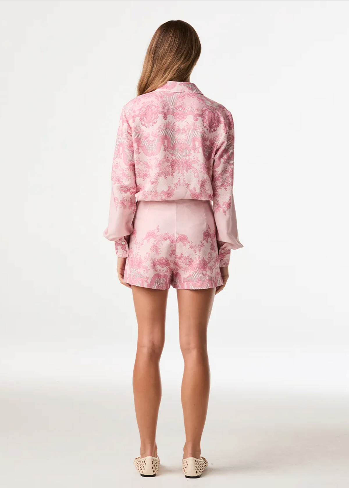 A woman viewed from behind wearing a pink and white patterned matching set consisting of a cropped long-sleeve jacket and shorts, paired with beige flat shoes against a plain white studio background.