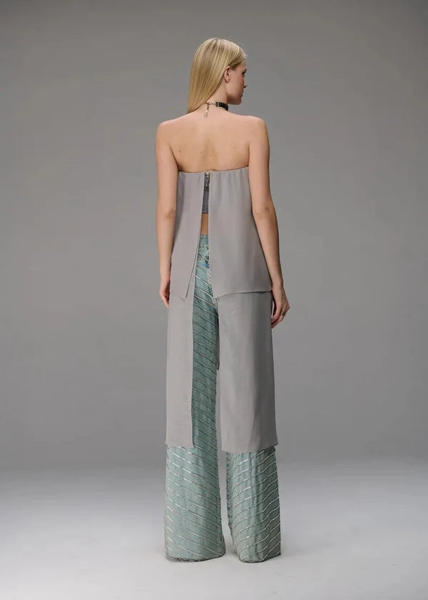 A model shown from the back wearing a strapless, wide-leg jumpsuit with a light gray upper section and textured sage green lower panels, set against a plain gray studio background.
