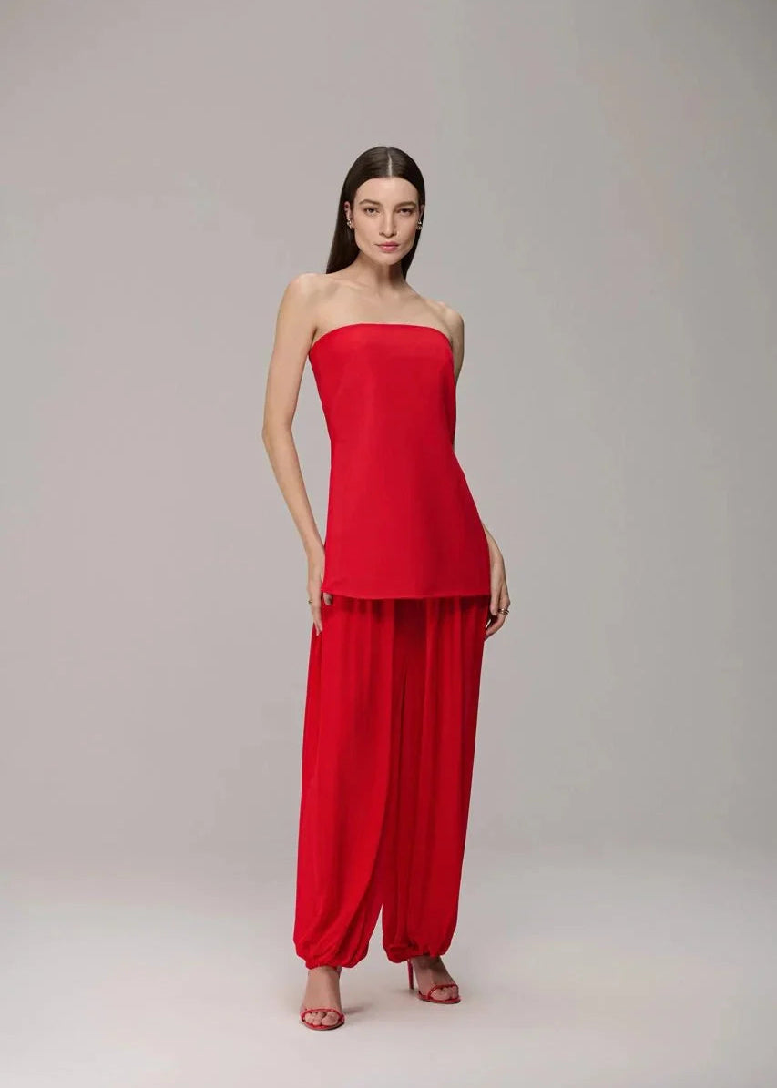 A woman modeling a bright red two-piece outfit consisting of a strapless bandeau-style top and matching loose harem pants, photographed against a plain light gray studio background.