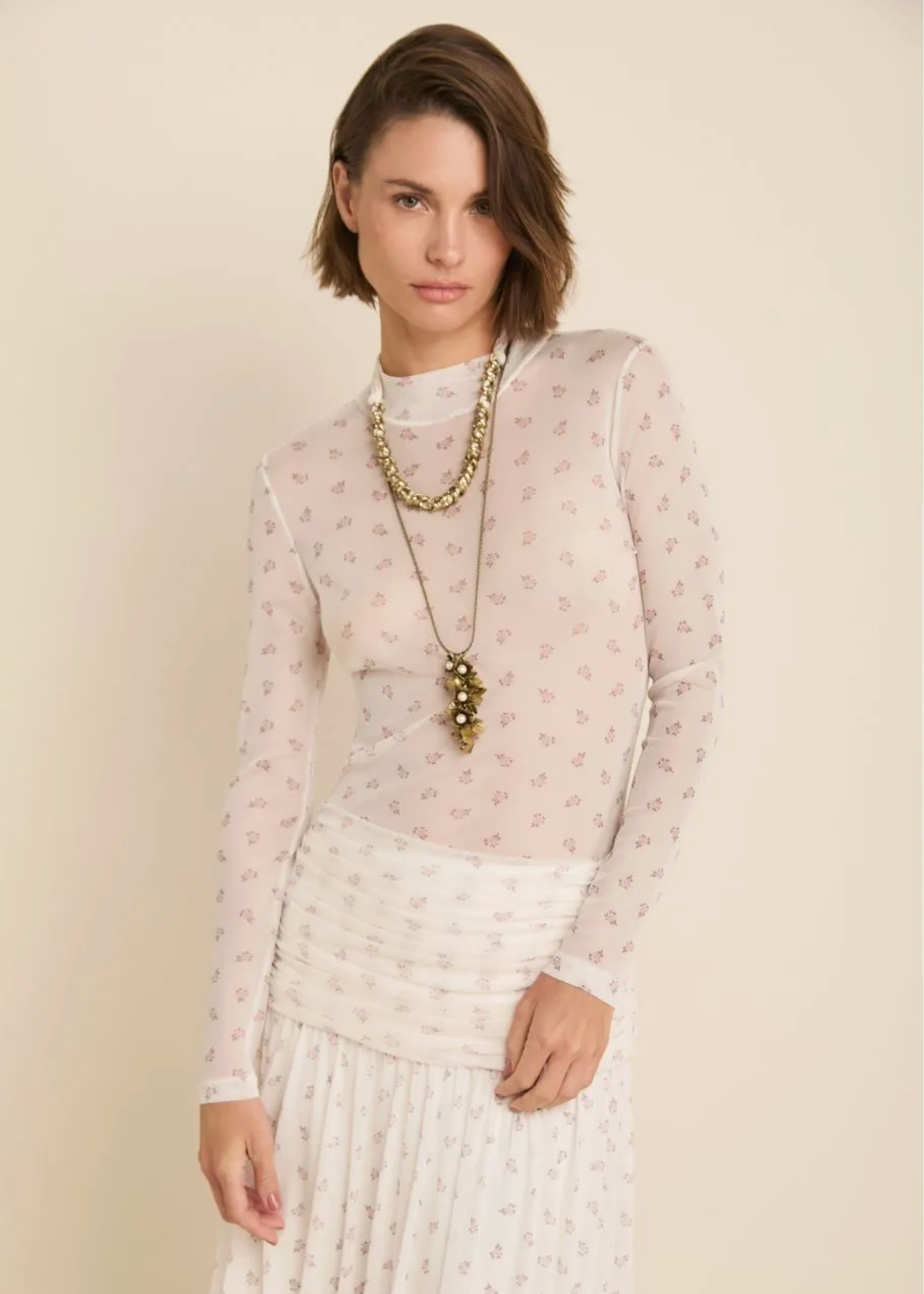 A woman modeling a sheer long-sleeve top in a pale blush pink and white floral pattern, paired with a matching white skirt and layered gold necklaces, set against a solid beige background.