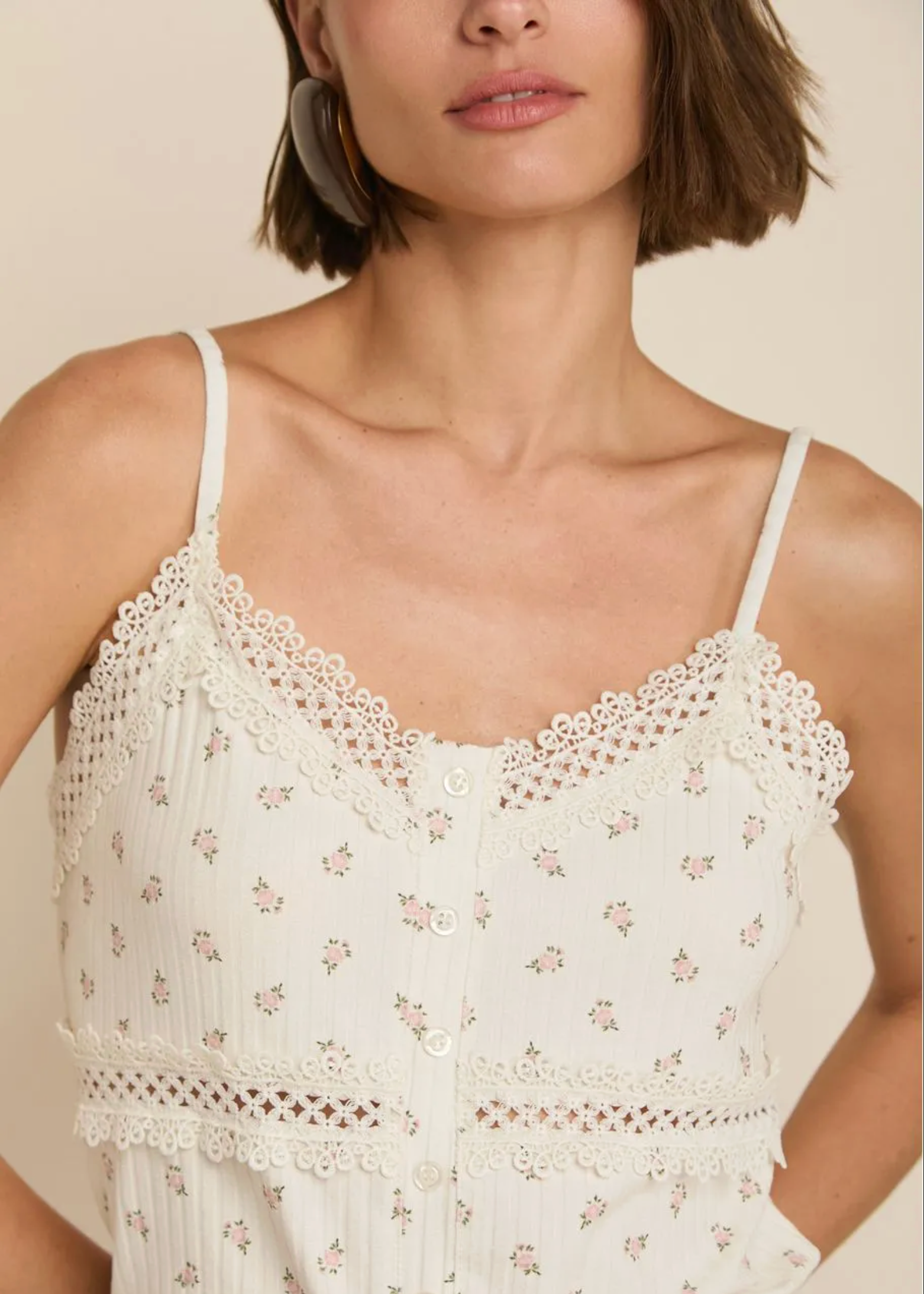 A close-up of a woman wearing a cream-colored floral lace camisole top with thin straps and a button-front design.