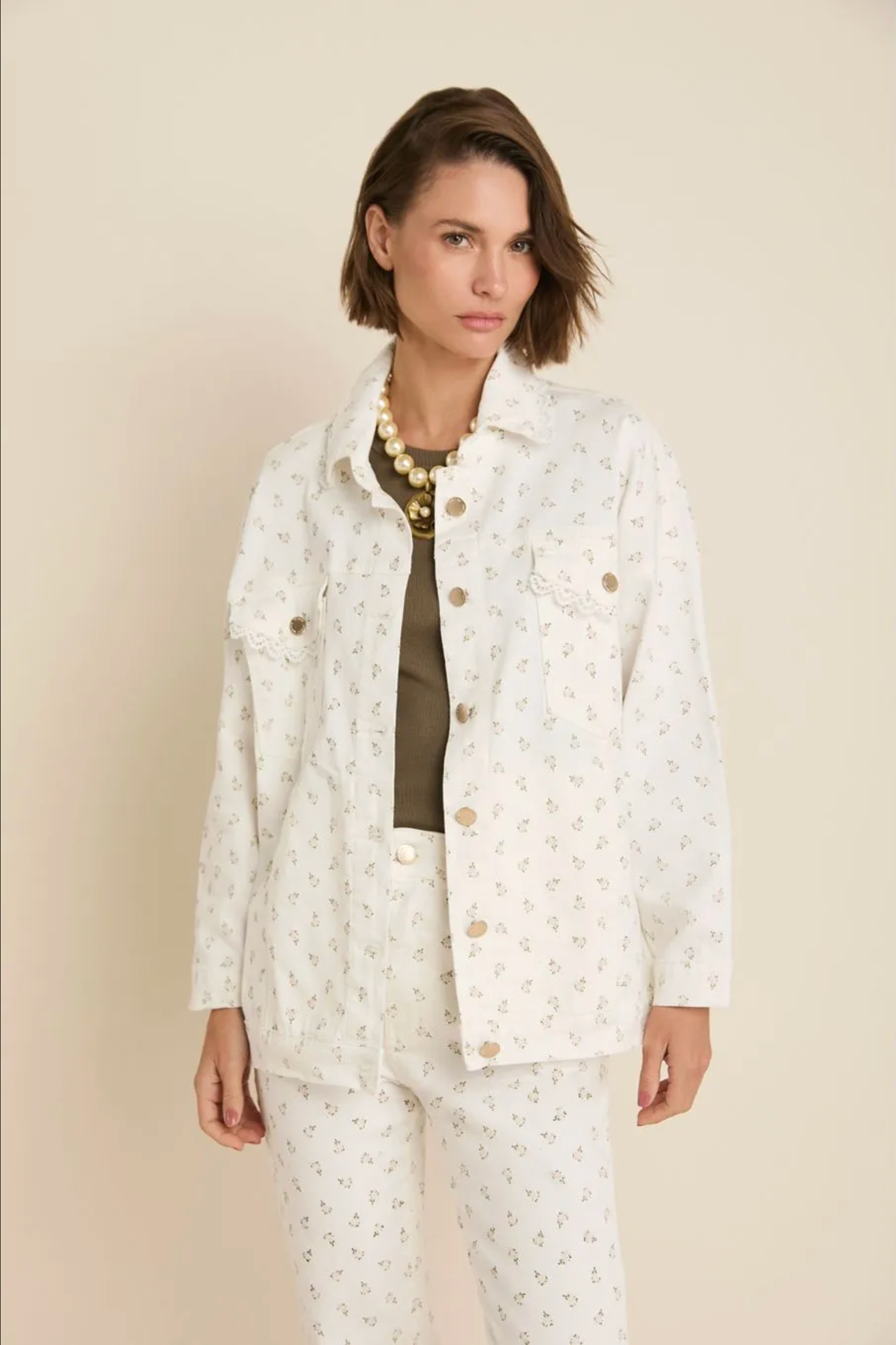 A woman modeling a matching white two-piece linen set with a small beige floral pattern, featuring a long-sleeve button-up shirt and coordinating pants, styled with a dark olive top and gold jewelry against a solid beige background.