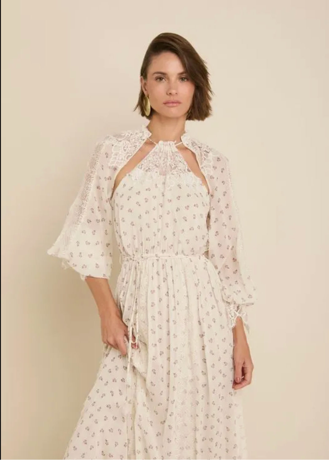 A woman modeling a long white maxi dress with a small all-over floral print, featuring ruffled elbow-length sleeves and a high neckline, posed against a solid cream studio background.