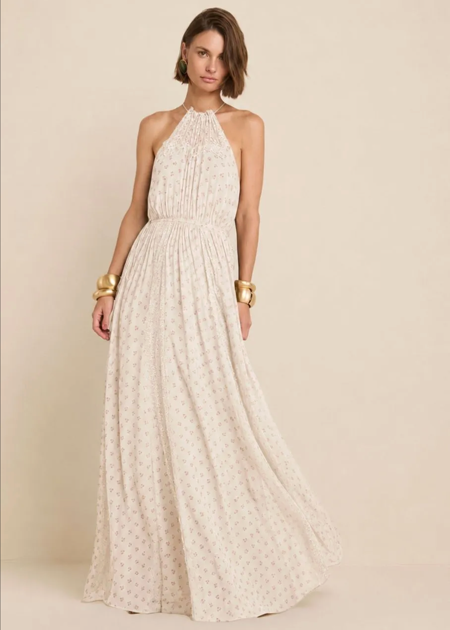 A woman modeling a sleeveless halter-neck maxi dress in a warm off-white or cream color with a small repeating floral print, set against a solid beige studio background.