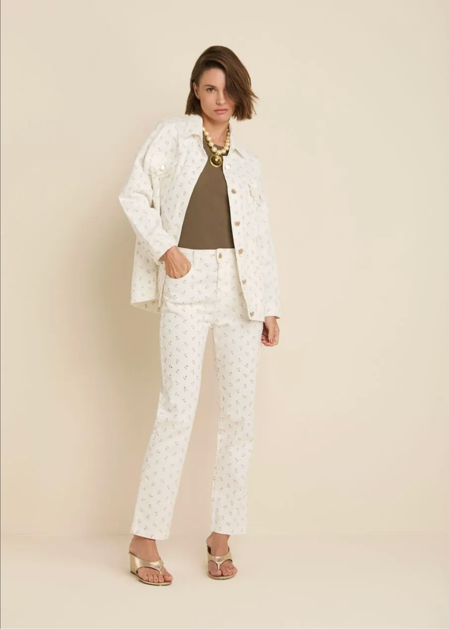 A model stands against a solid beige studio background wearing a matching white patterned jacket and trousers set, styled with a brown top, a chunky gold necklace, and gold sandals.
