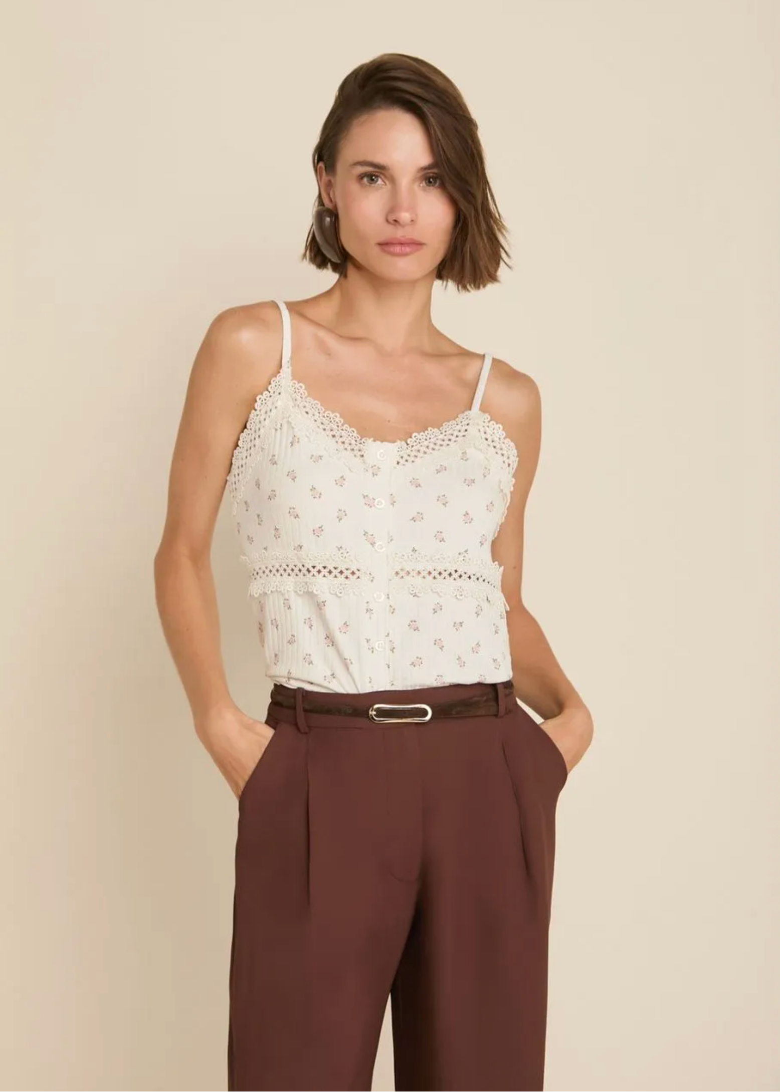 A woman modeling a white eyelet camisole top with delicate floral embroidery and thin straps, paired with brown trousers and a dark belt, against a solid beige studio background.