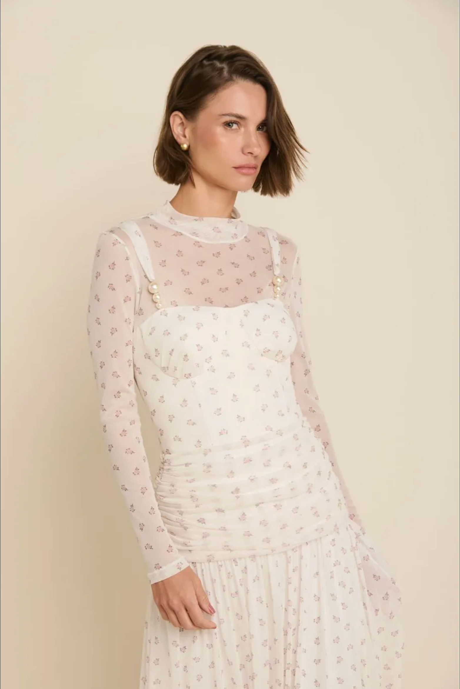 A woman wearing a sheer long-sleeve mesh top with a small pink floral print over a white camisole dress with matching skirt, posed against a solid light beige background.