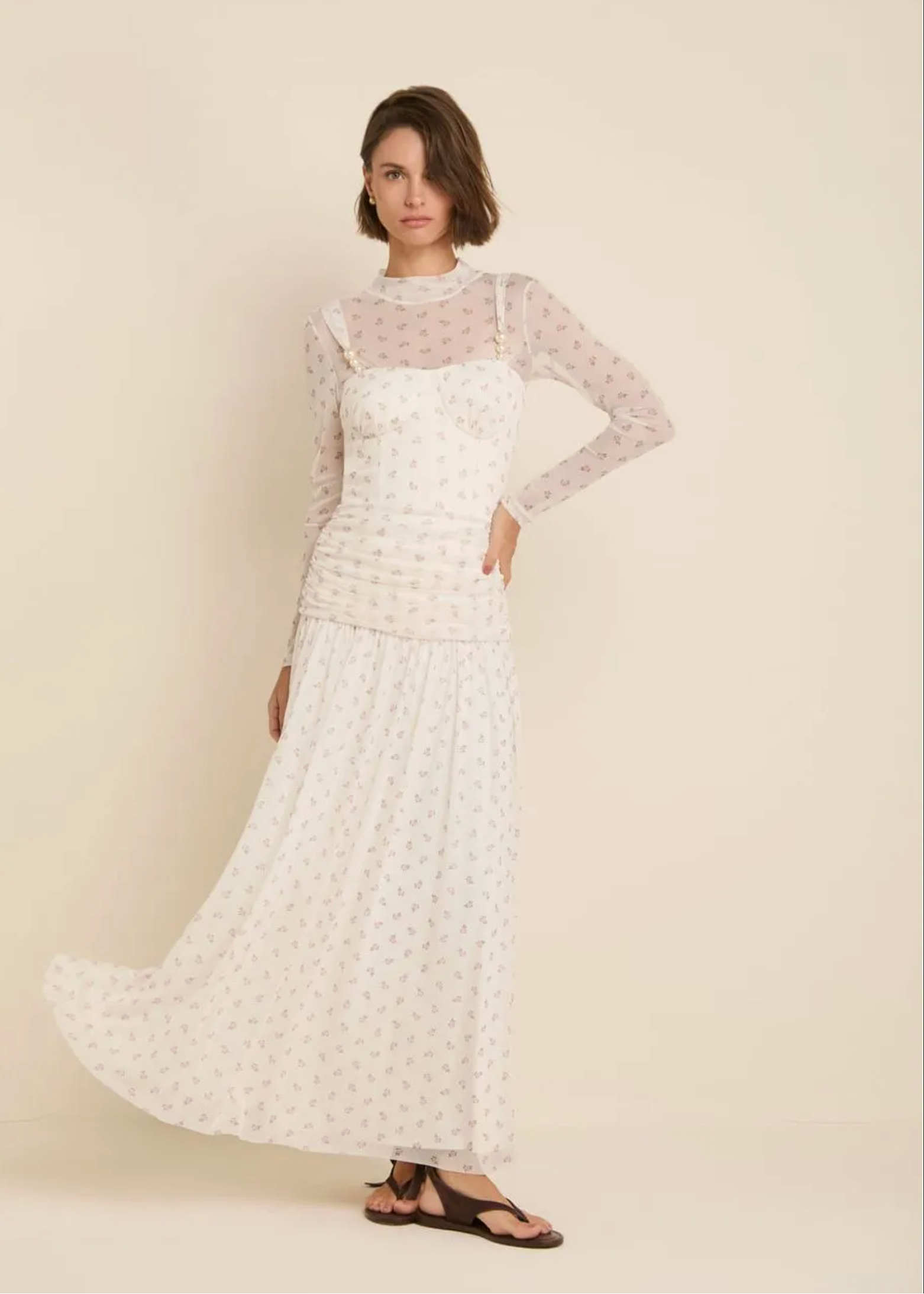A woman modeling a long-sleeve white maxi dress with a delicate pink floral print, featuring a sheer mesh overlay at the neck and shoulders, set against a solid cream background.