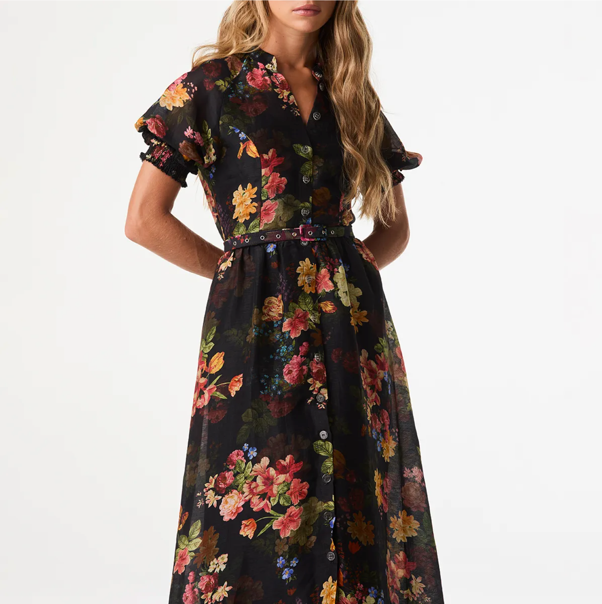 A woman wearing a black midi dress with a vibrant multicolor floral print, featuring short puff sleeves, a button-down front, and a matching fabric tie belt at the waist, set against a plain white studio background.