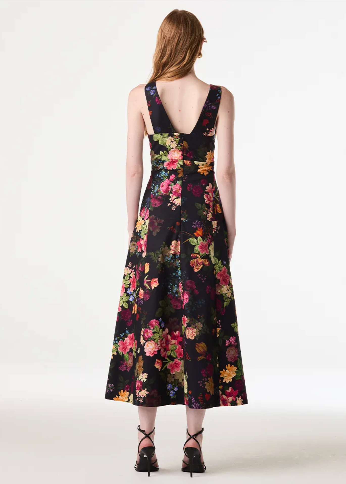 A woman stands with her back to the camera wearing a sleeveless black midi dress with a vibrant multicolor floral print and a deep V-back. She is wearing black lace-up high heels against a plain white studio background.