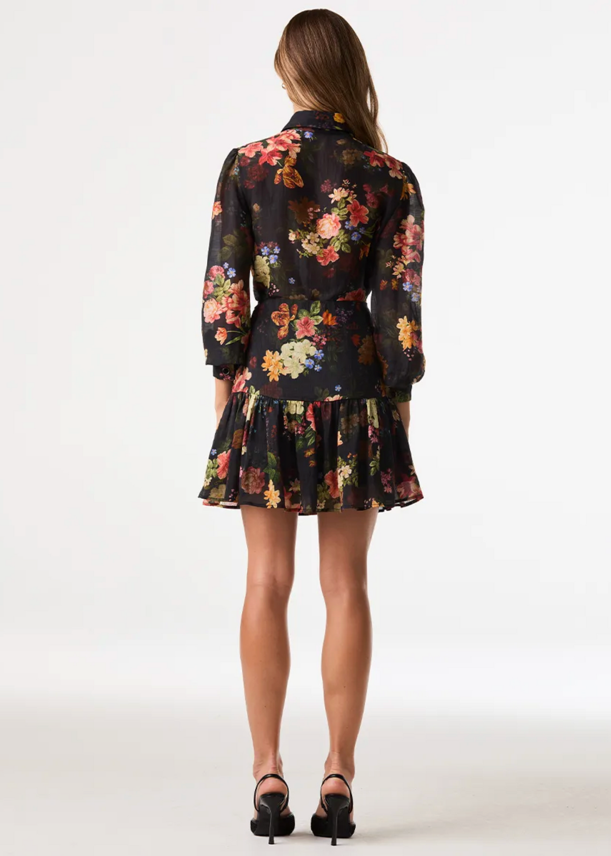 A woman is shown from the back wearing a black long-sleeve mini dress with a vibrant multicolor floral pattern, paired with black high heels, against a plain white studio background.