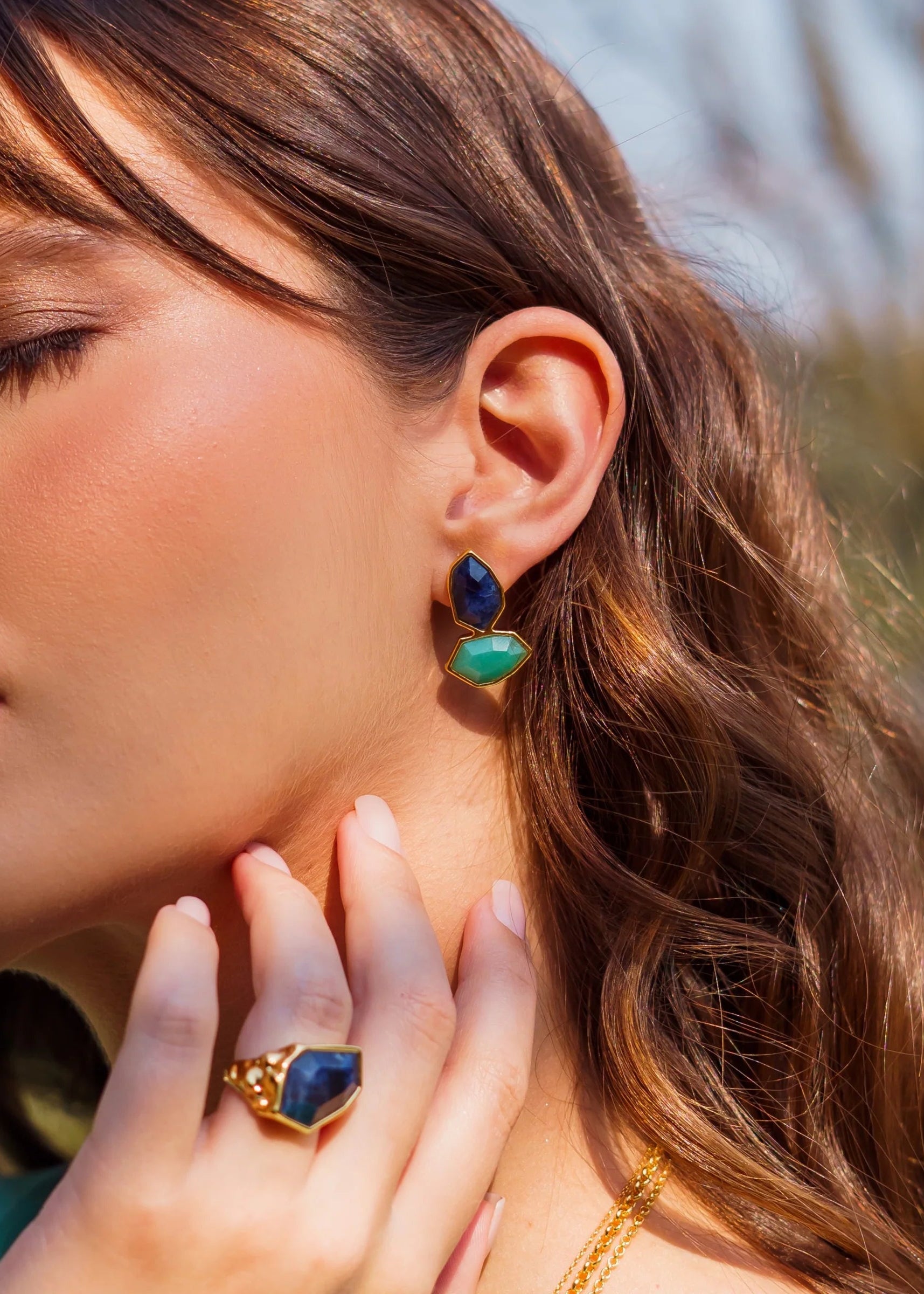 Close-up of a woman wearing blue and green earrings and a matching ring outdoors.