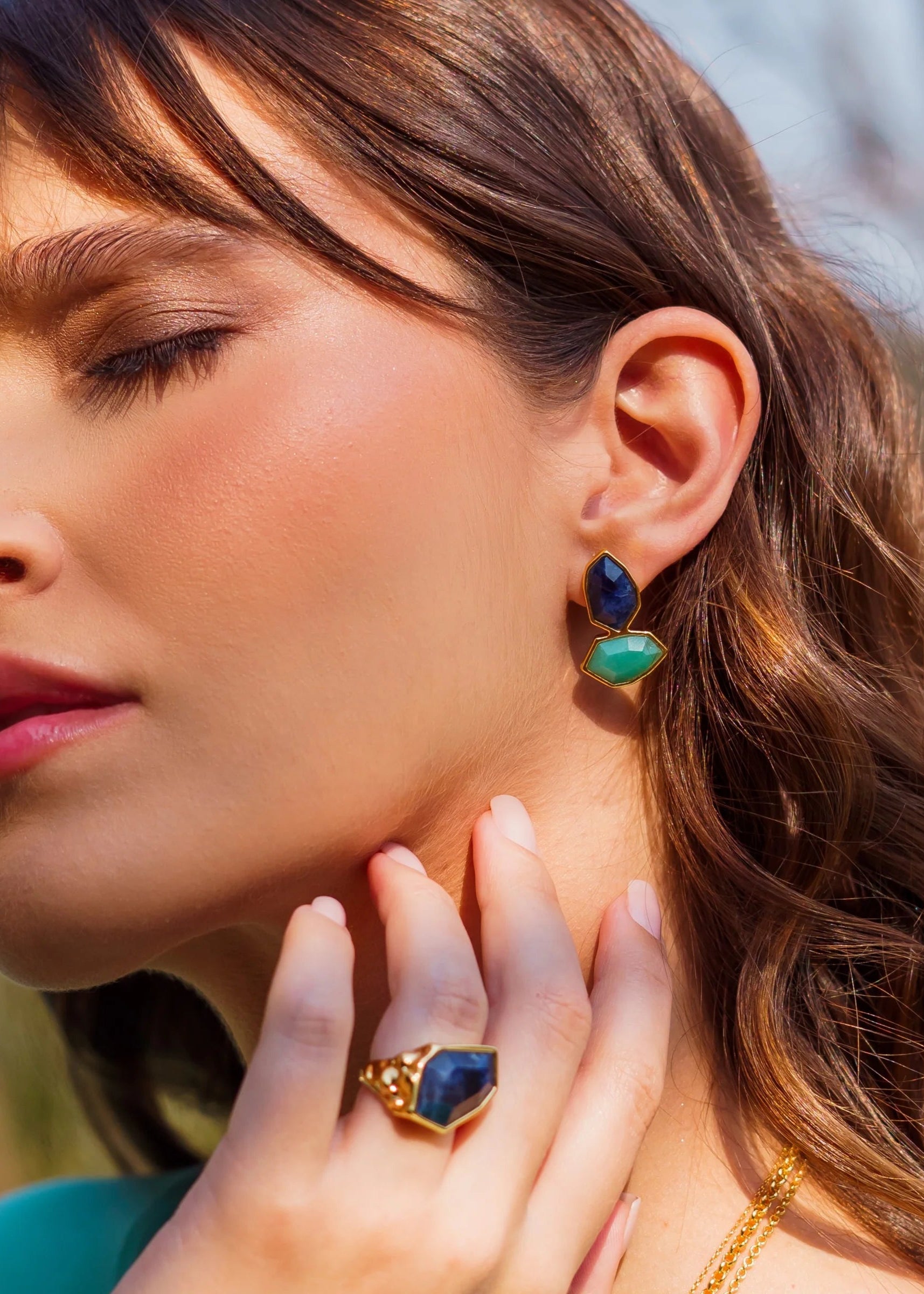 Close-up of a woman wearing blue and green earrings and a matching ring outdoors.
