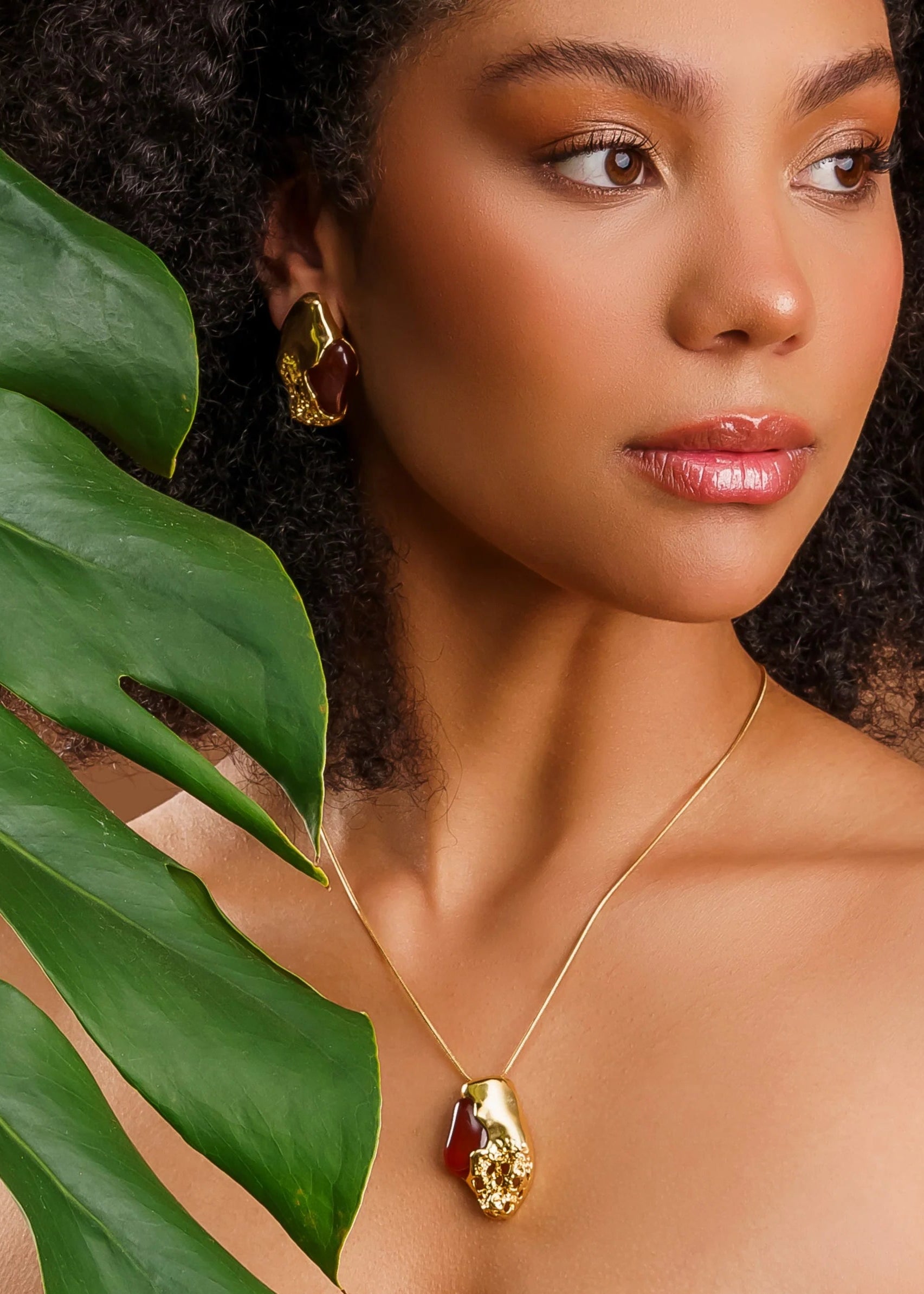 Woman wearing a gold necklace with a red gemstone, surrounded by green leaves.