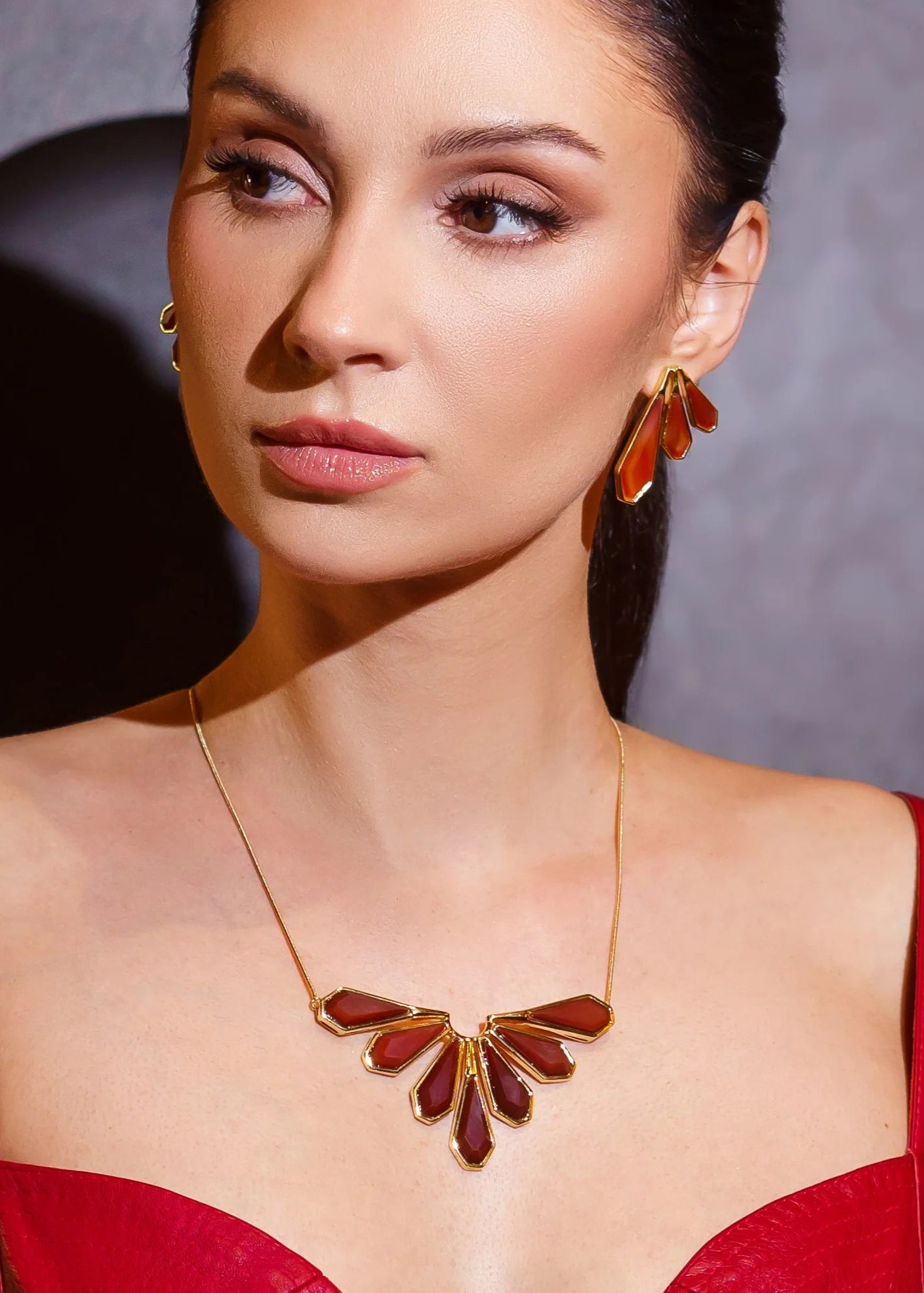 Woman wearing a gold necklace and earrings with a red dress against a gray background