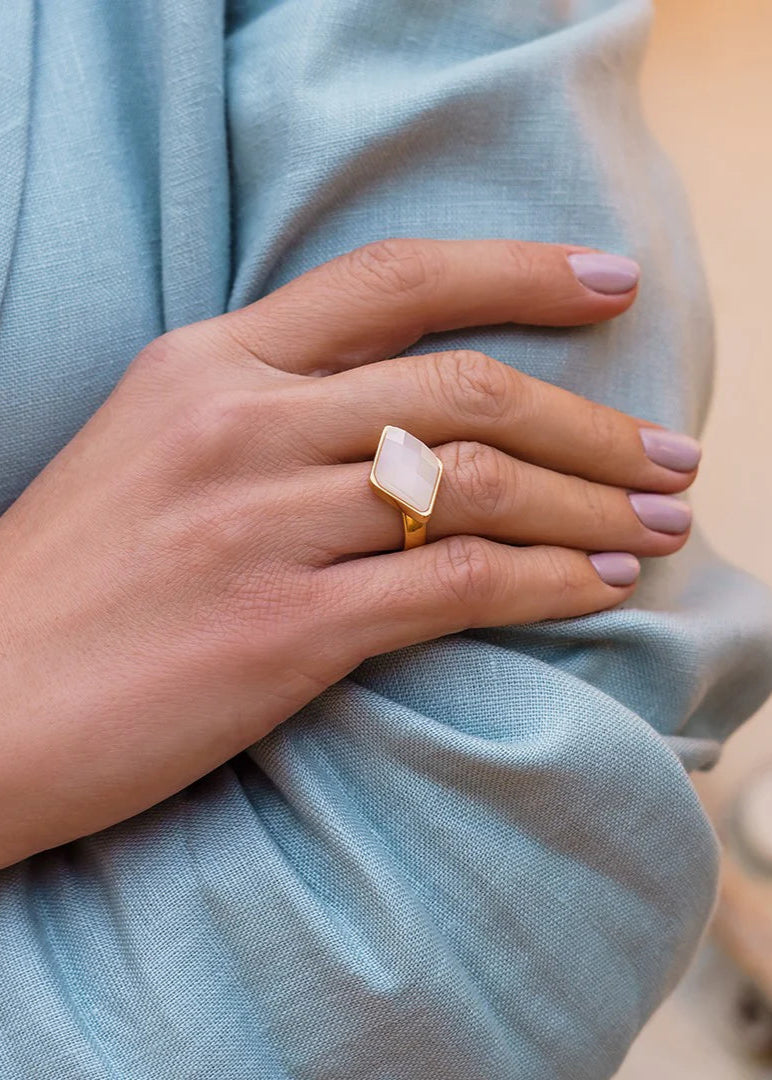 Hand wearing a gold ring with a white stone on a light blue fabric background