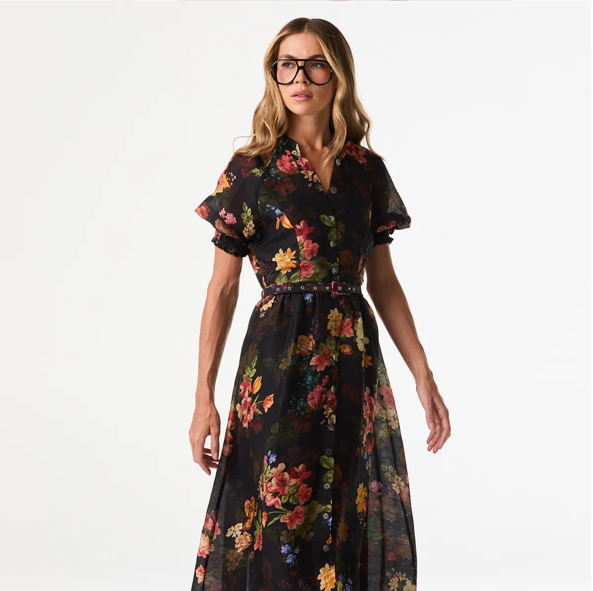 A woman modeling a black midi wrap dress with a vibrant multicolor floral print, sheer flutter sleeves, and a matching waist tie, set against a plain white studio background.