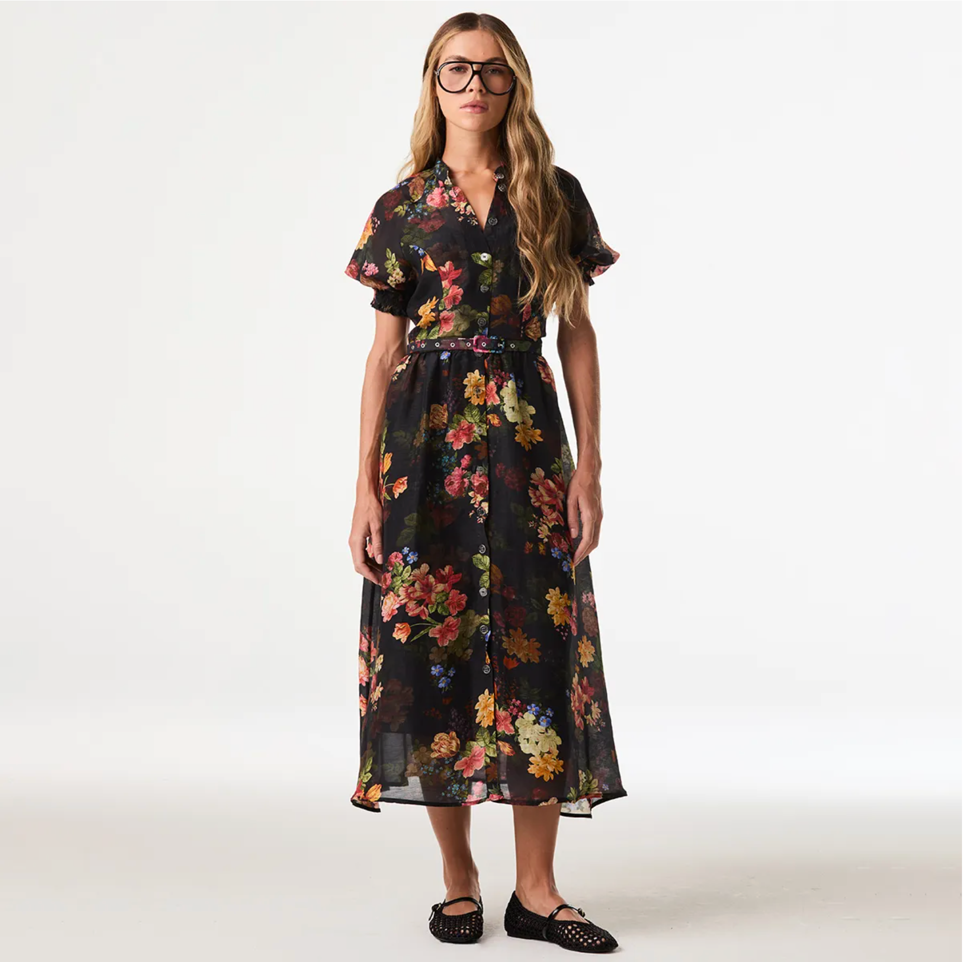 A woman modeling a black midi dress with a vibrant multicolor floral print, featuring short flutter sleeves, a collared neckline, and a matching fabric tie belt, set against a plain white studio background.