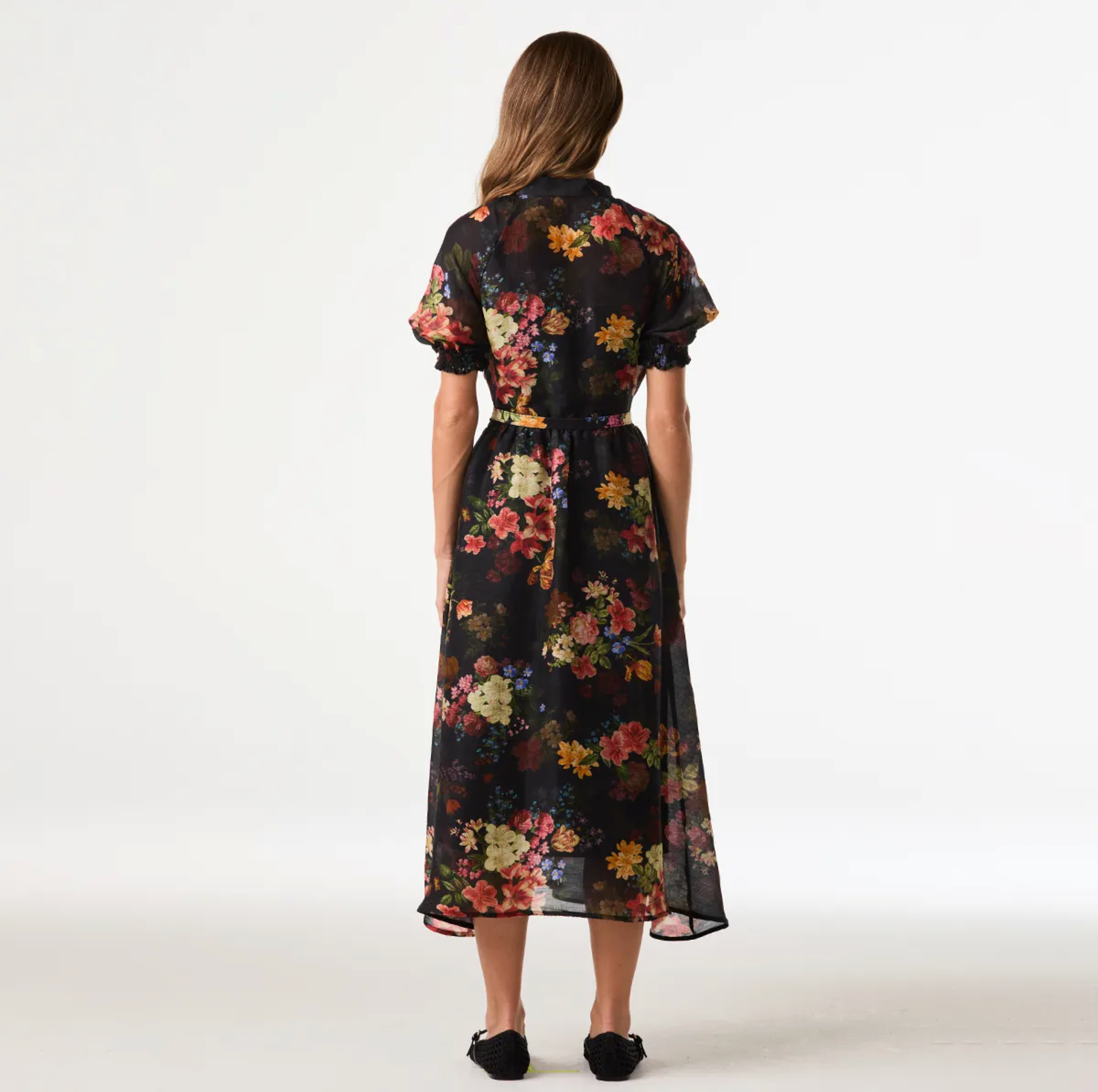 A woman viewed from the back wearing a short-sleeve midi dress with a black base and a colorful floral print, paired with black flats, against a plain white studio background.
