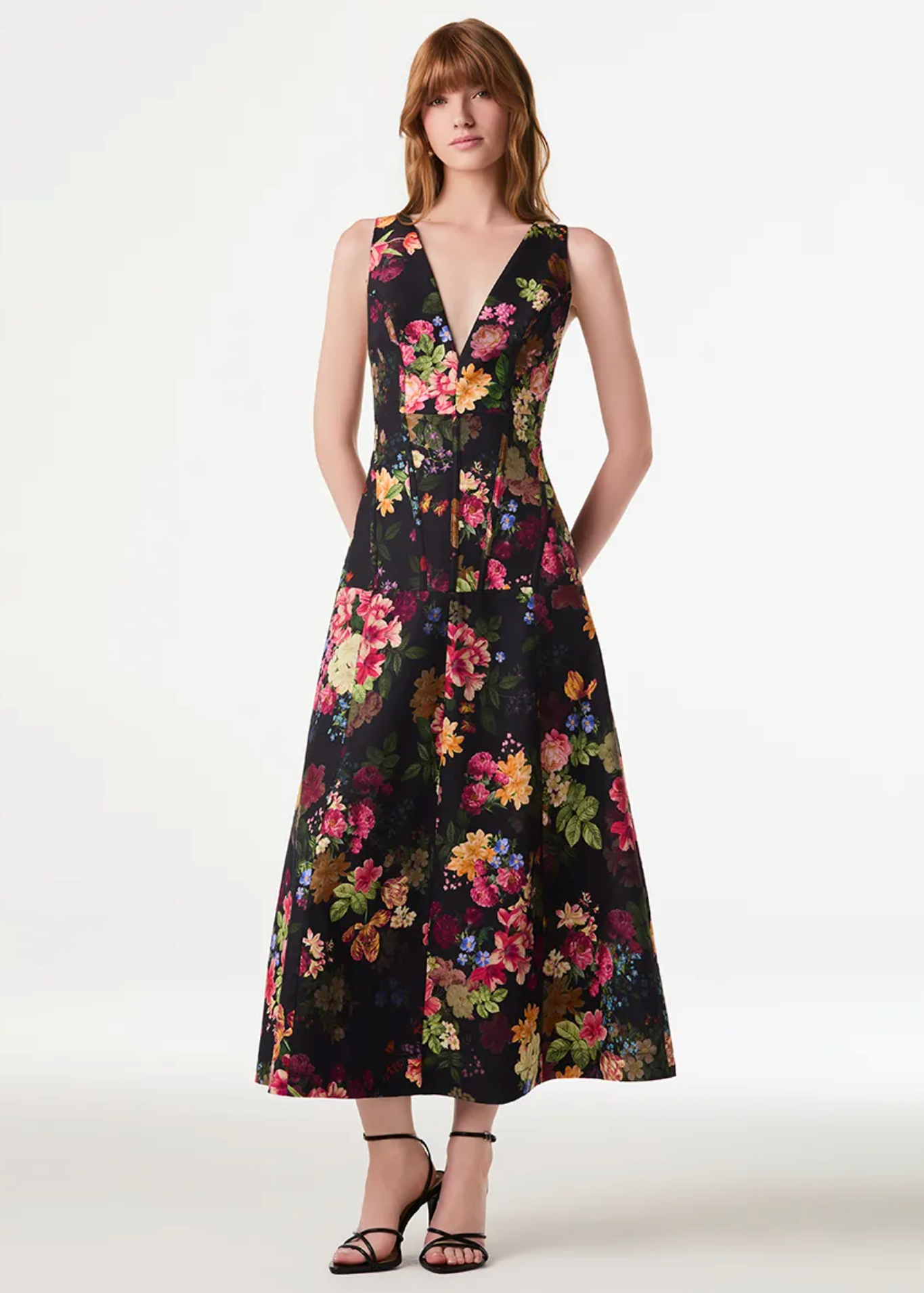 A woman modeling a sleeveless black midi dress with a vibrant multicolor floral print, featuring a deep V-neckline and a flared skirt, set against a plain white studio background.
