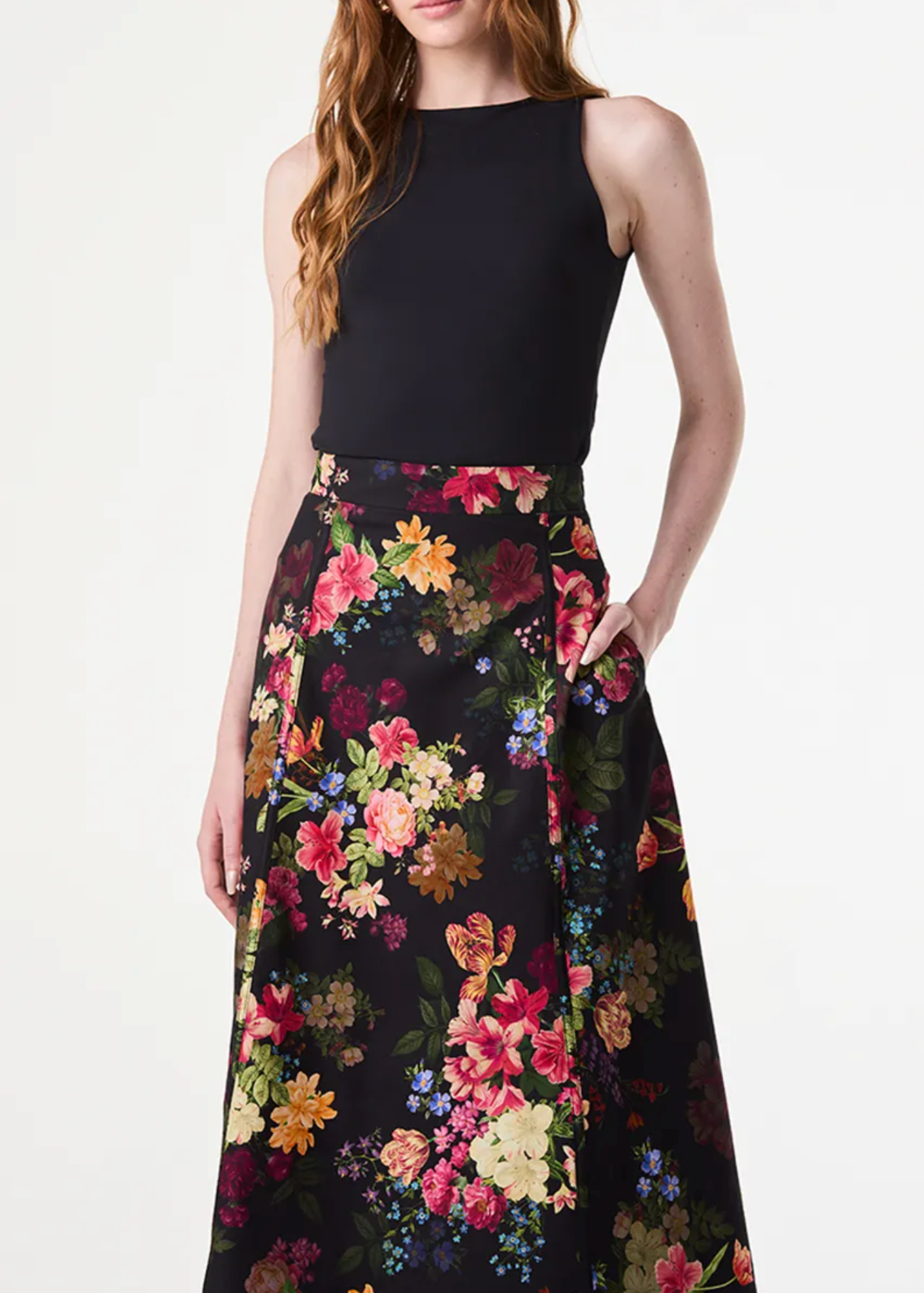 A model wearing a sleeveless black top and a high-waisted black midi skirt with a vibrant multicolor floral print, set against a plain light gray studio background.