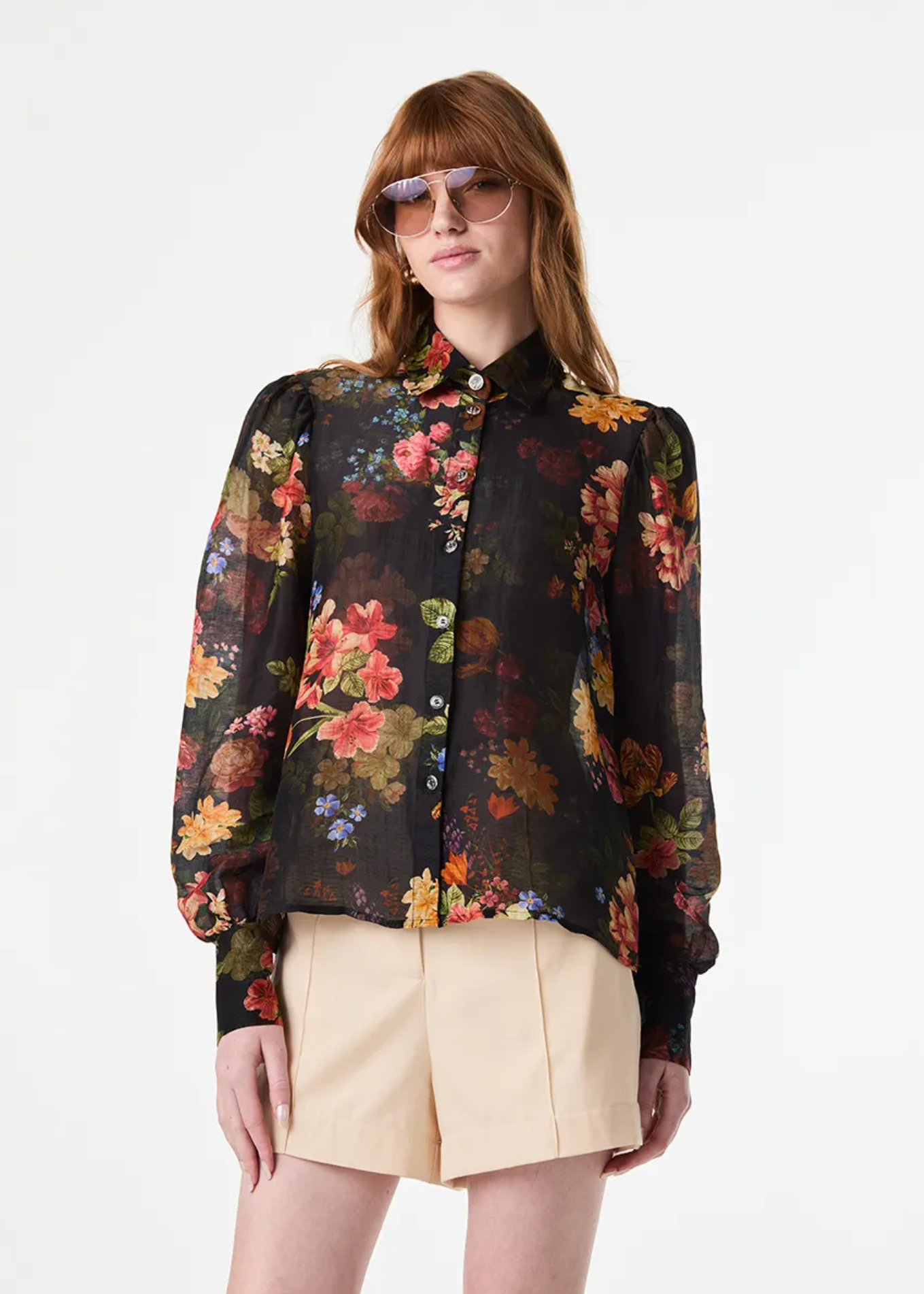 A woman modeling a sheer black long-sleeve blouse with a vibrant multicolor floral print, paired with beige shorts, against a plain white studio background.