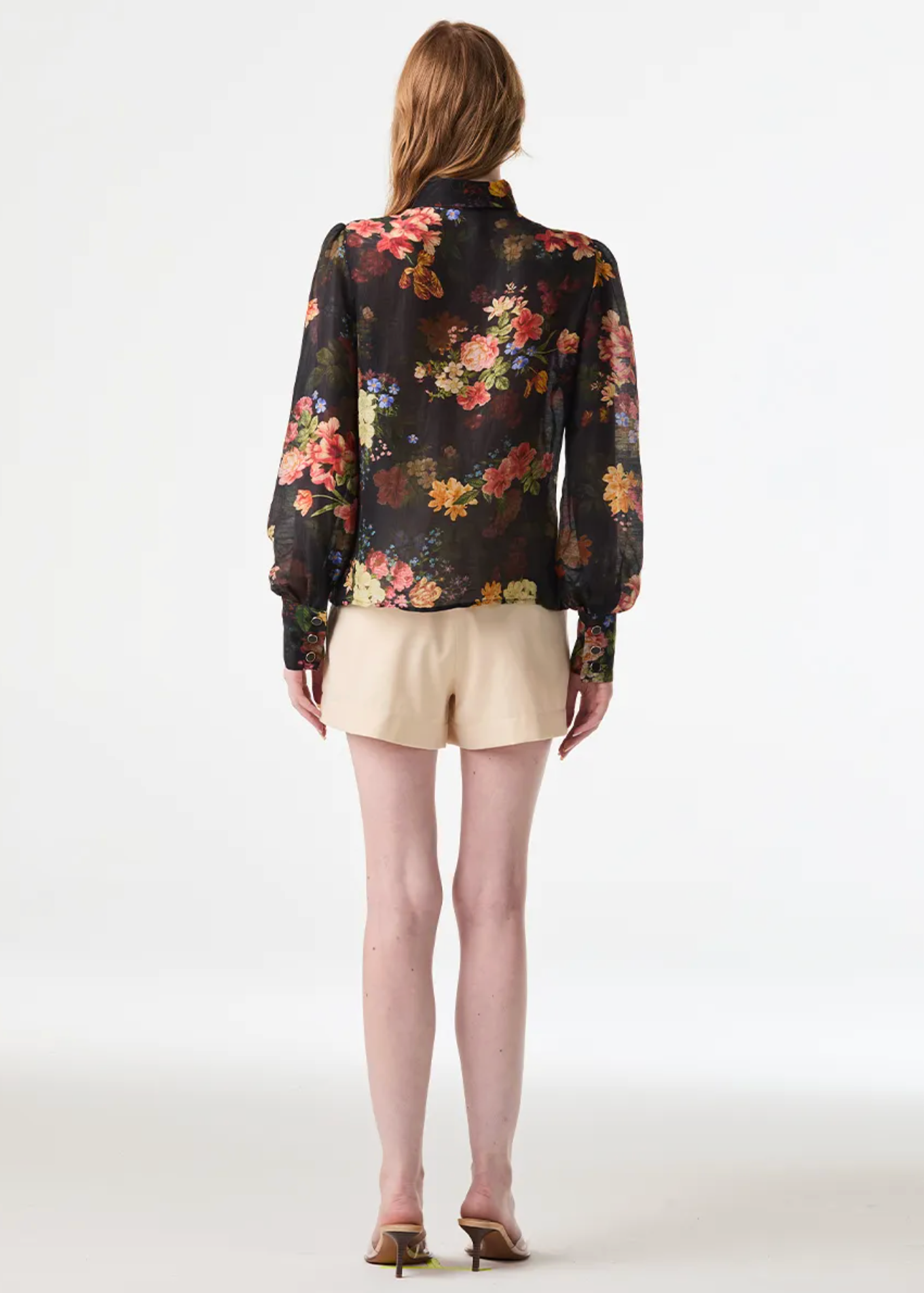 A woman is shown from the back wearing a black long-sleeve blouse with a vibrant multicolor floral print, paired with beige shorts and brown heels, against a plain white studio background.