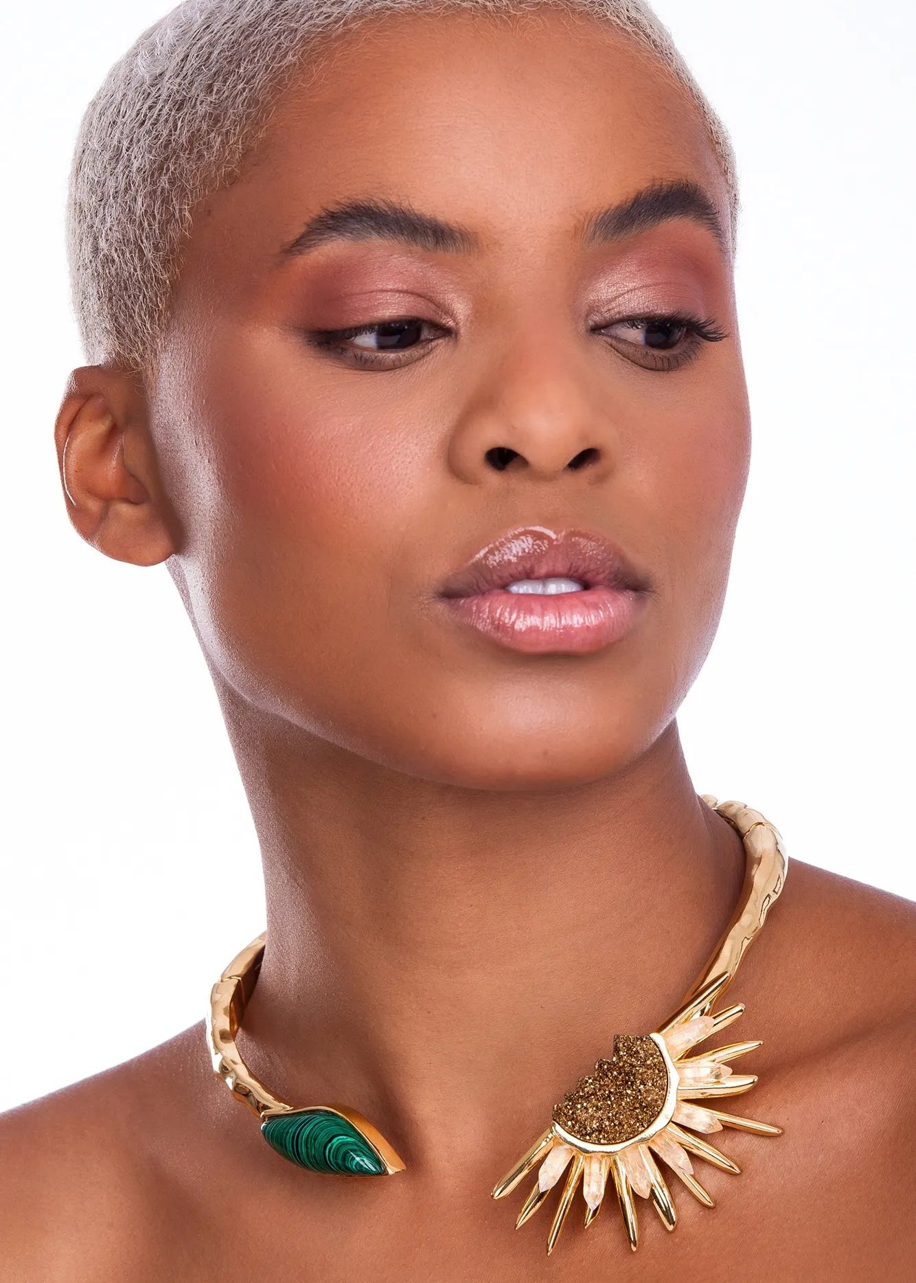 A close-up of a model wearing a gold-toned statement necklace with a sunburst design and a green teardrop pendant, set against a plain white background.
