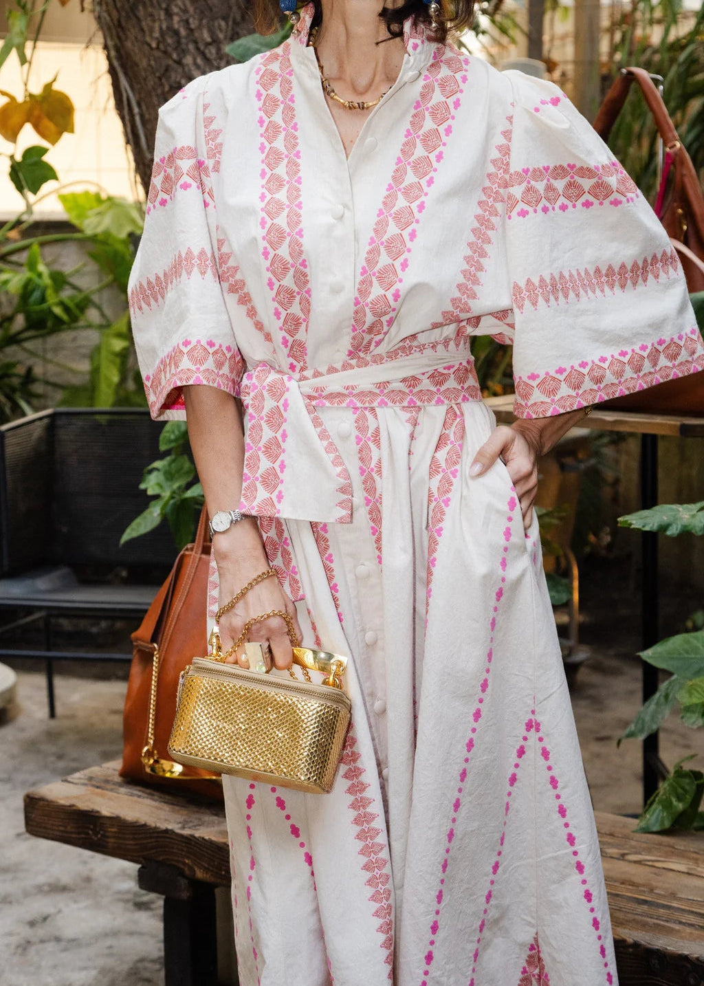 Woman wearing a white dress with pink patterns, holding a gold handbag, in an outdoor setting.