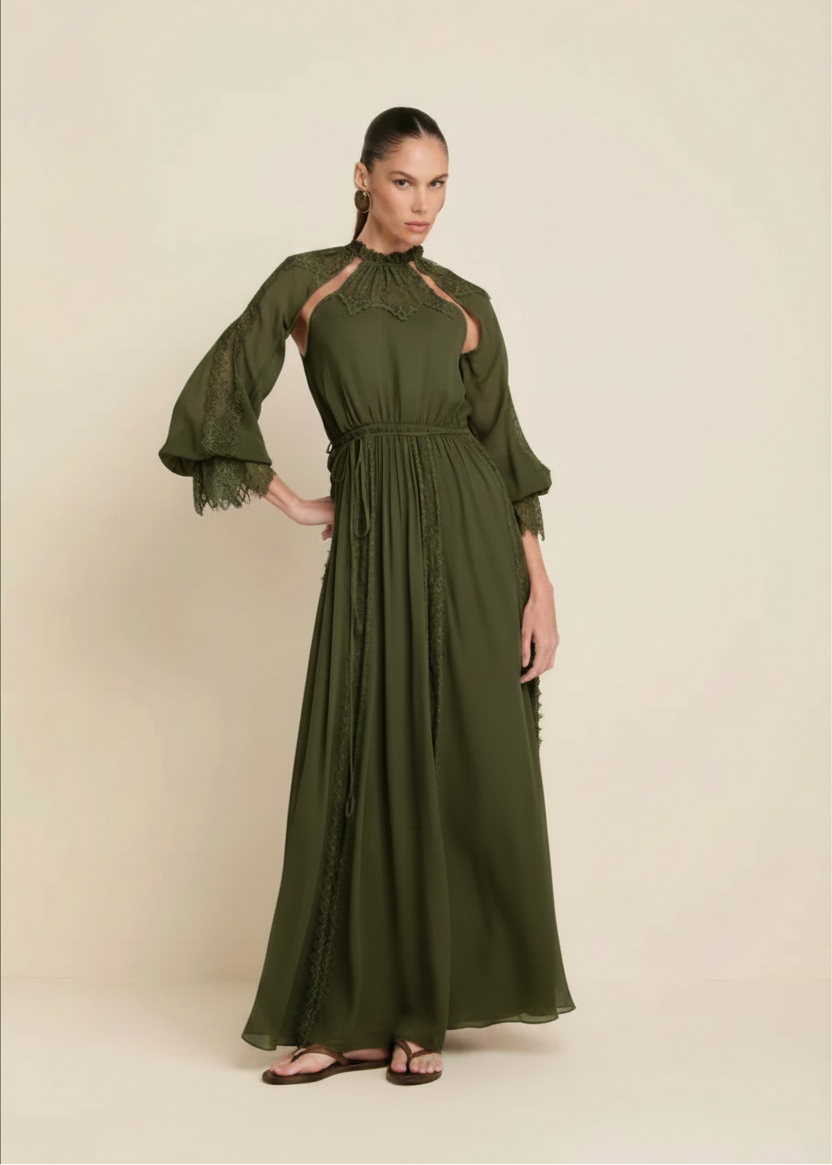 A woman modeling a long brown maxi dress with sheer lace details, high neck, and flared sleeves, standing against a plain beige studio background.