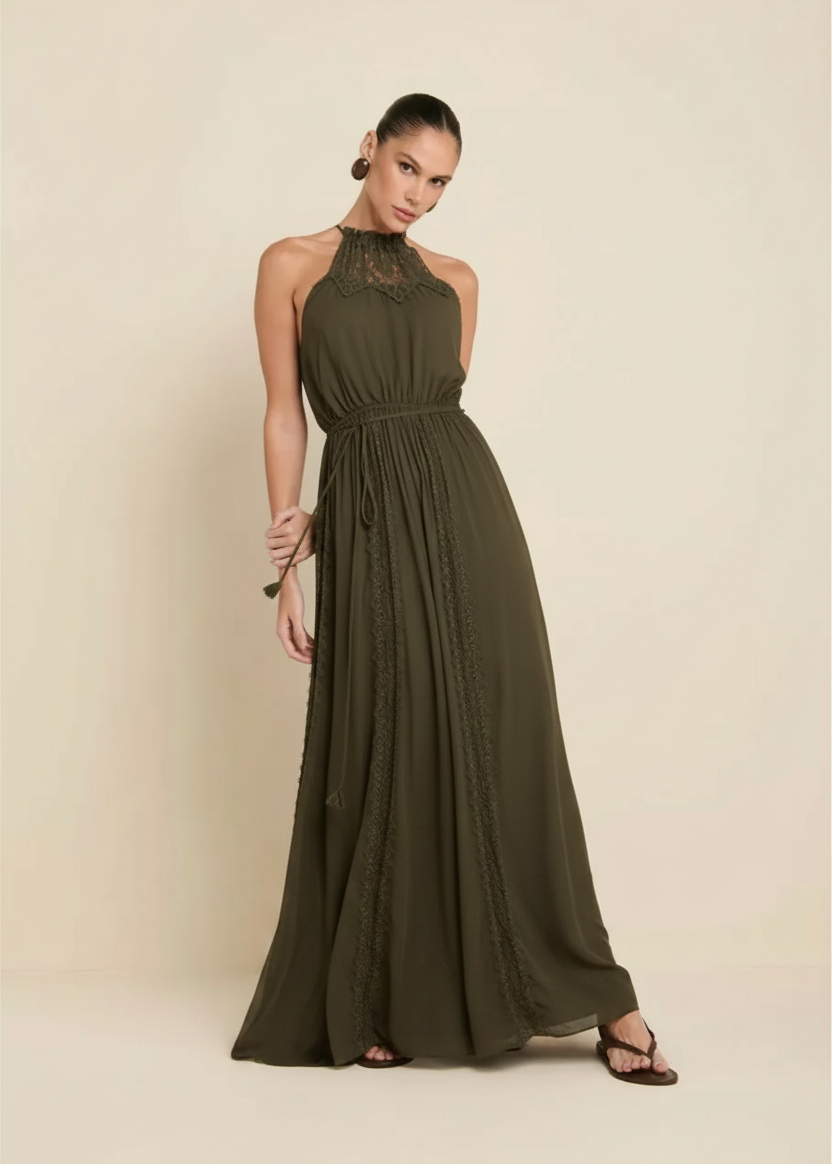 A woman modeling a sleeveless, high-neck, floor-length brown dress with lace detailing at the neckline and along the sides, set against a solid beige studio background.